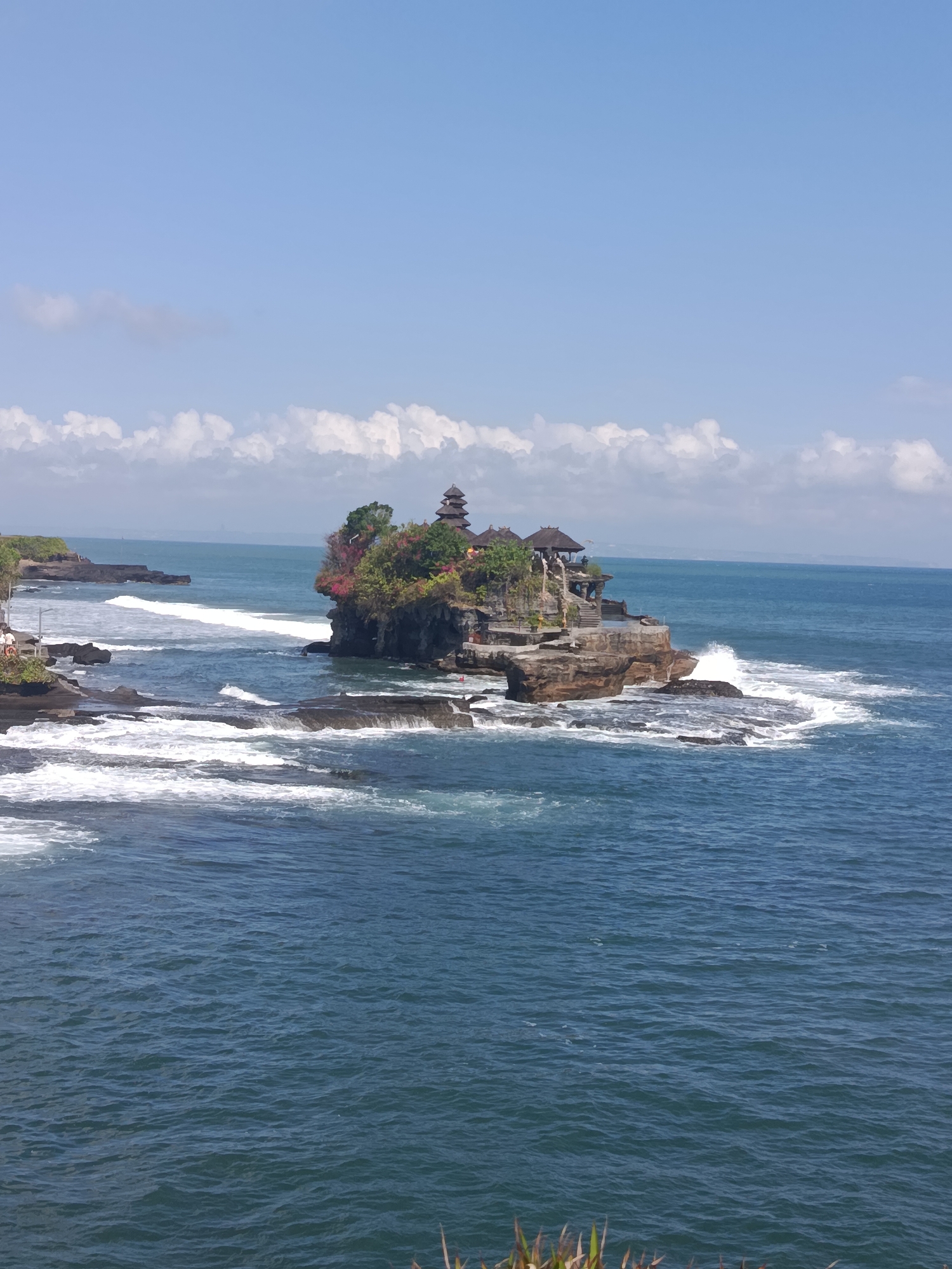 The image depicts Tanah Lot Temple, a famous sea temple located in Beraban, Bali, Indonesia. The temple is situated on a rock formation offshore, and waves crash against the base of the rock. The water is a deep blue, and the sky is clear with a few white clouds. The temple itself is composed of several tiered structures with traditional Balinese architecture. Green vegetation and colorful flowers adorn the top of the rock. In the foreground, there are rocky shorelines with smaller waves breaking. The overall scene is serene and picturesque, capturing the iconic beauty of the location.