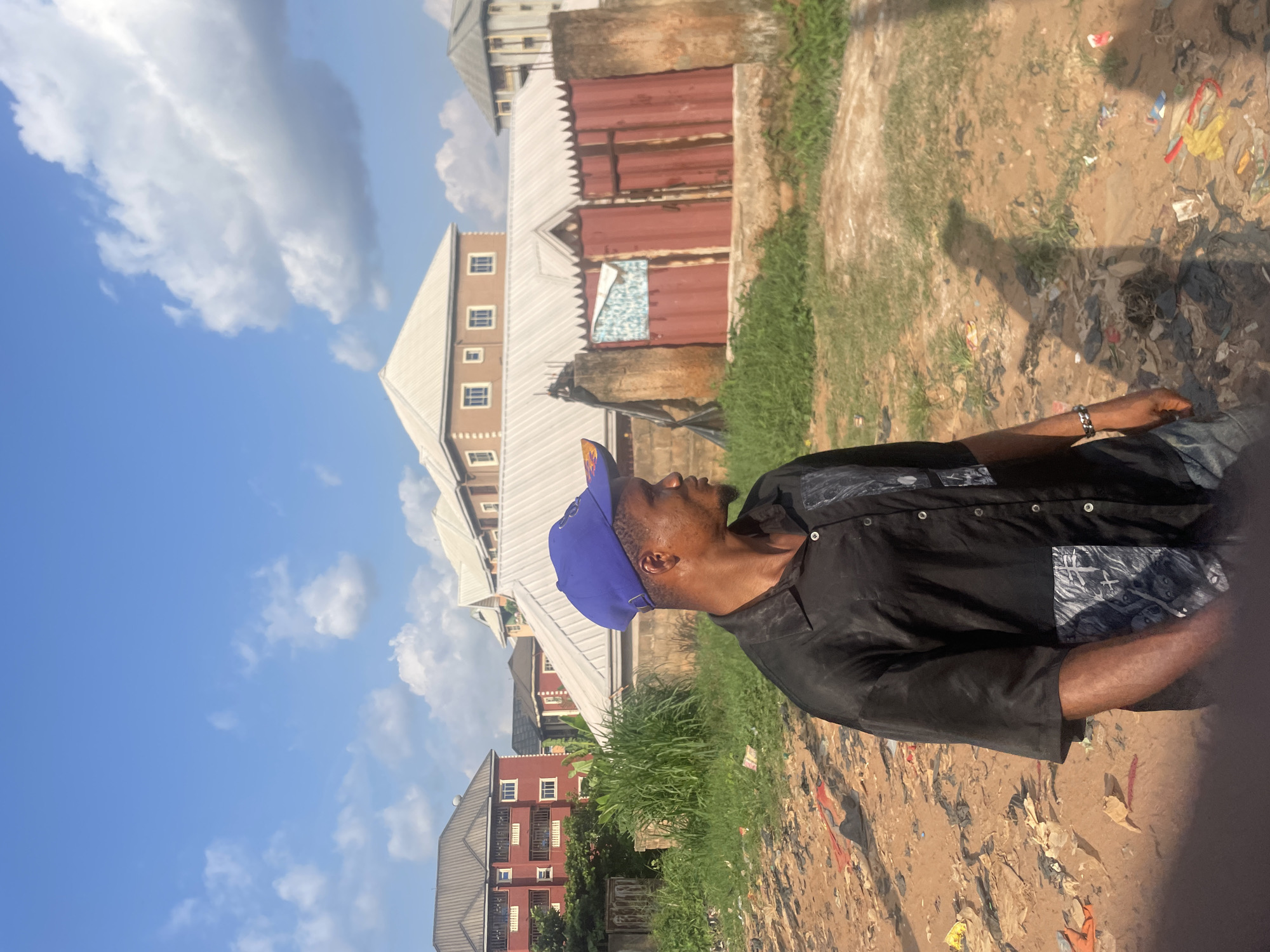 The image captures a candid moment of a man standing outdoors in Nkpor, Nigeria, under a bright, sunny sky. The man, seen from the side, is dark-skinned and wears a blue baseball cap and a black short-sleeved collared shirt with light-colored patterns on the chest and shoulders. A silver bracelet adorns his right wrist. He is looking slightly upwards and to his left.
The background features several buildings under a clear blue sky with scattered white cumulus clouds. These include multi-story structures with white pitched roofs and numerous windows, one appearing reddish-brown. A reddish-brown corrugated metal gate is visible directly behind the man. The ground is uneven, composed mostly of dry dirt, interspersed with patches of green grass and littered with plastic, paper, and fabric scraps. The bright lighting indicates it is daytime, likely late morning or early afternoon. No specific activity or strong emotions are discernible, and no visible text is present.