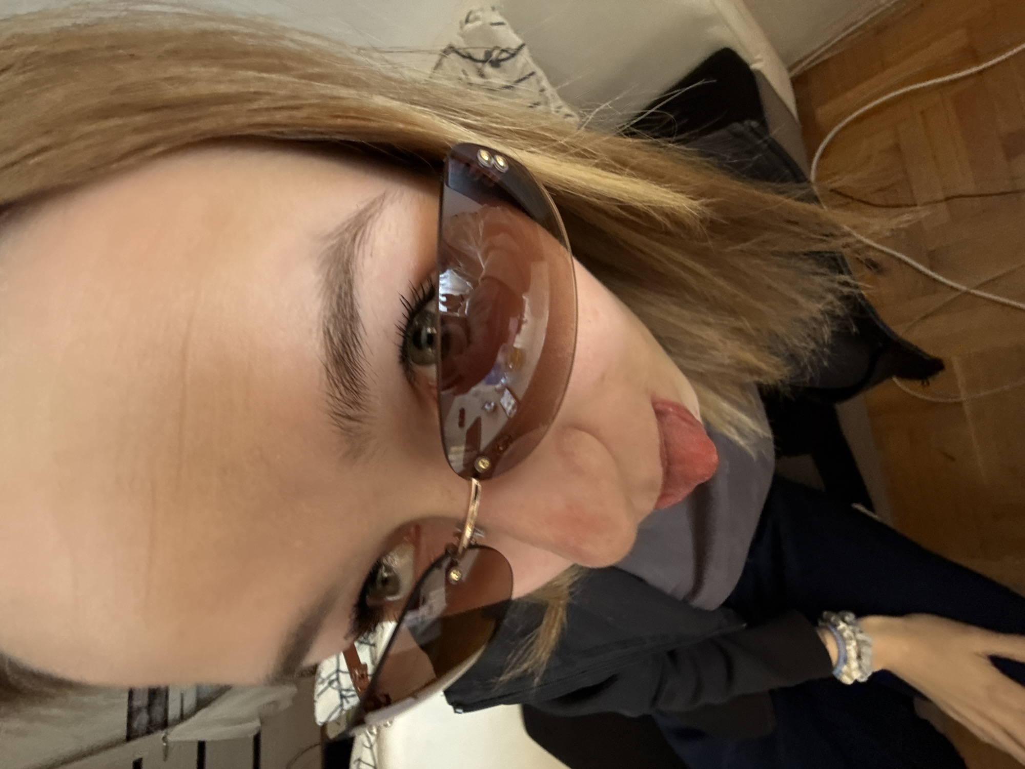 A close-up, angled shot captures a person, likely female, indoors. The individual has light brown/blonde hair and is wearing brown-tinted sunglasses. Their left eye, visible beneath the sunglasses, appears green or hazel with dark eyelashes. A notable detail is the person playfully sticking out their tongue towards the viewer. The sunglasses reflect internal room elements, possibly furniture or light sources.
The person is dressed in a dark-colored jacket or top, with what seems to be a grey garment visible underneath. On their left wrist, a multi-strand bracelet adorned with blue, white, and silver beads or charms is visible. The background shows hints of an indoor setting: light-colored wooden floorboards with a white cable running across them, and what might be a patterned textile or cushion behind the person's head. The image has a casual and lighthearted feel, indicated by the playful facial expression. There is no visible text in the image. The time of day cannot be determined, but the scene is adequately lit. While the context states Kragujevac, Serbia, no specific visual cues within the image itself confirm this location.