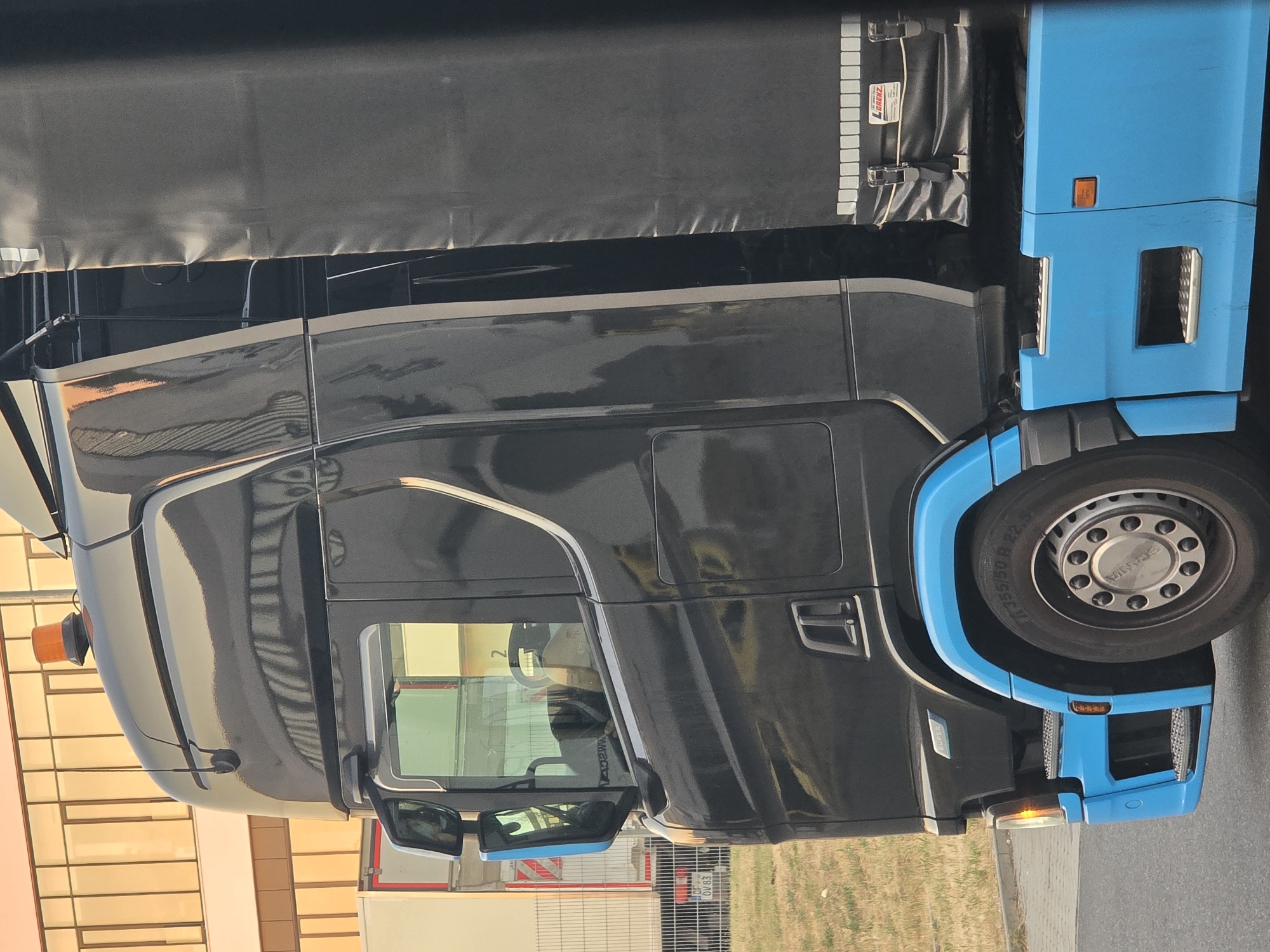 The image shows the right side of a black semi-truck parked on a paved surface, possibly a street or parking lot, with a grassy area and a building in the background. The truck has bright blue accents on the lower bodywork and wheel arches. The truck has a large side mirror with blue housing, and a clear window showing the interior of the cab. A black tarp covers the cargo area of the truck. There is a yellow flashing light on the roof of the cab. A license plate reading "OF OV 83" is visible on a white trailer behind the truck. The building in the background has a golden, reflective facade with a grid pattern. The scene appears to be in Kelsterbach, Germany, based on the context provided. There are no people visible in the image.