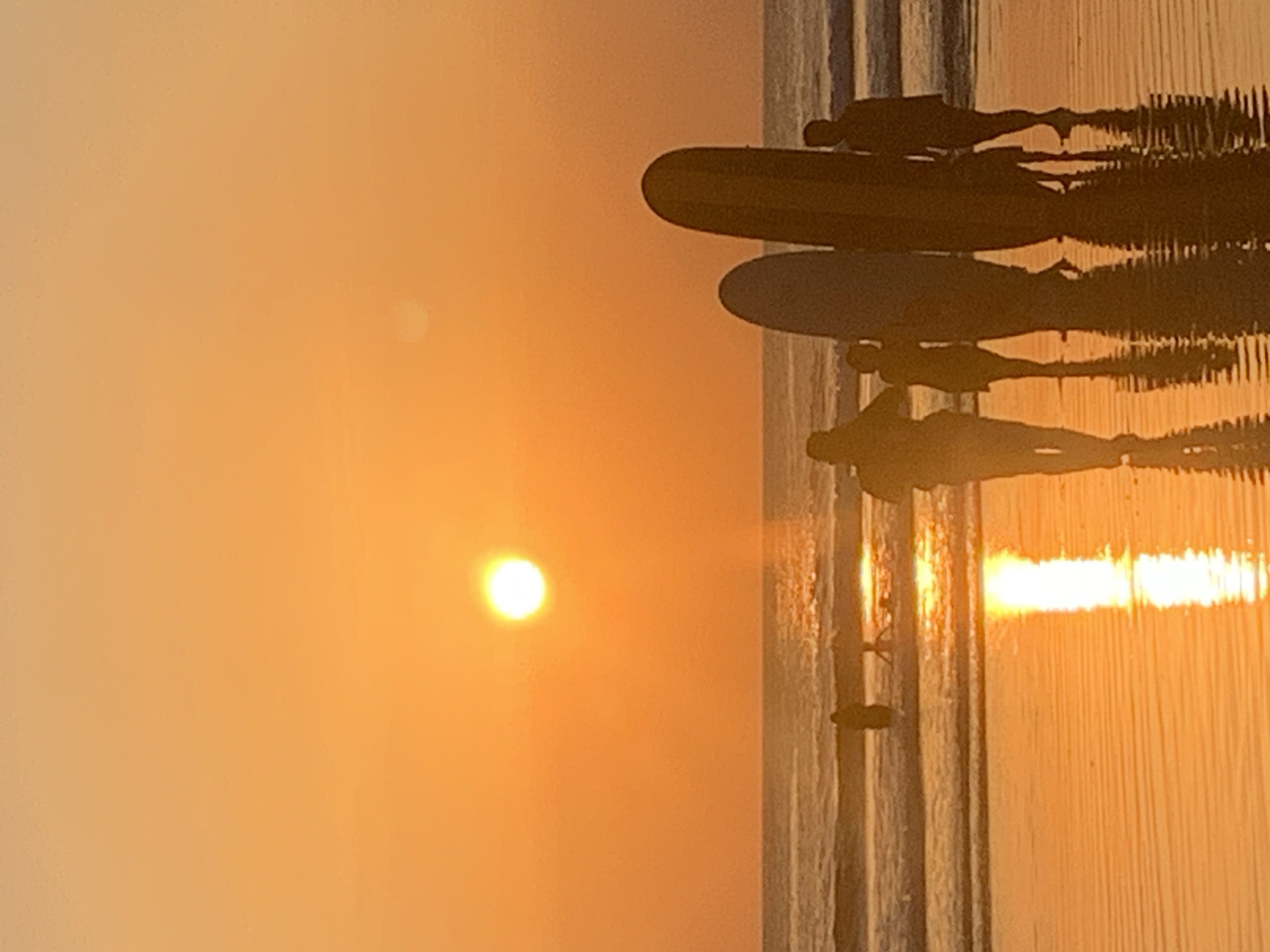 The image captures a serene sunset scene at Kuta Beach, Bali, Indonesia. The sun, a bright, glowing orb, is setting over the ocean, casting a warm, orange hue across the sky and reflecting vibrantly on the wet sand. Several people, silhouetted against the sunset, are standing on the beach, some with surfboards. The waves are gentle, creating a peaceful atmosphere. The overall impression is one of tranquility and natural beauty.