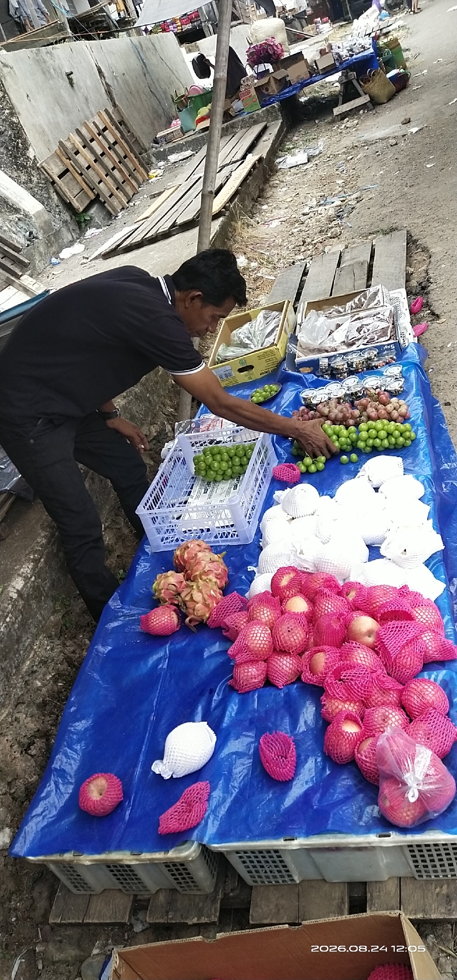 A man in a black shirt and black pants is seen at a street market stall in Tamiang Layang, Indonesia. He is interacting with various fruits displayed on a blue tarp, which covers several white plastic crates. The fruits include green grapes in a white plastic basket, pink and white dragon fruit, and apples wrapped in pink netting. Behind the man, there are boxes, bags, and more fruits. The stall is set up on a dirt and gravel surface, with wooden planks and pallets visible in the background. The environment appears to be an outdoor market, possibly in a less developed area of the city, given the rustic setup and the debris on the ground. The overall scene depicts a typical street vendor operation.