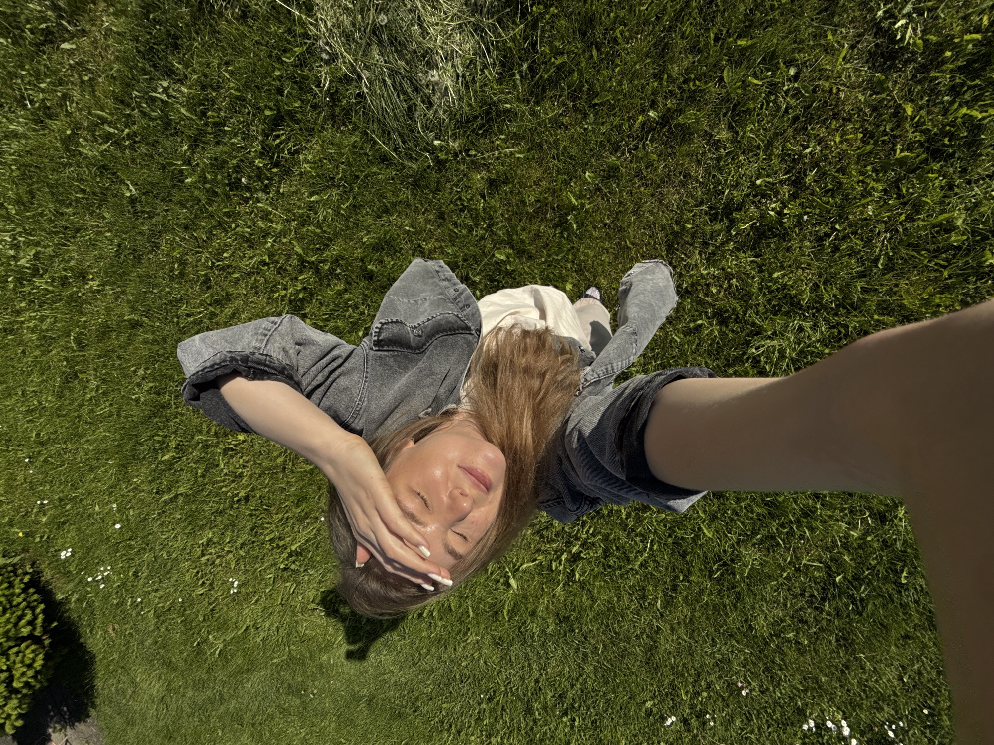 An overhead, high-angle shot captures a young woman relaxing outdoors on a sunny day. She is positioned centrally, lying or reclining on a vibrant green lawn. The woman has long, light brown hair and is wearing a grey denim jacket over lighter-colored clothing. Her eyes are closed, and her left hand is raised to her forehead, shading her face from the bright sunlight, conveying a sense of relaxation and contentment. Her fingernails are painted white.
The scene is dominated by lush green grass, with scattered small white flowers, possibly daisies, adding detail to the foreground. In the upper portion of the frame, some taller, drier grass is visible. A dark green bush or shrub appears in the lower left corner. The strong, direct sunlight casts clear shadows, indicating it is daytime, likely midday or early afternoon. The overall atmosphere is peaceful and serene, with the woman enjoying the natural environment. No text is visible in the image. The location context for this image is Красный Маяк, Russia.
