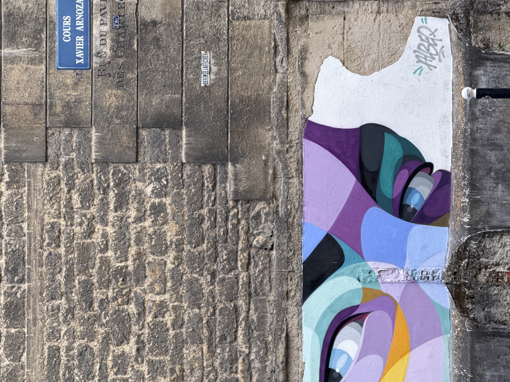 This image depicts a street art mural on a textured stone wall in Bordeaux, France. The mural features an abstract design with bold, swirling shapes in shades of purple, blue, teal, and yellow, with black and white accents. The artwork appears to be a close-up of eyes or facial features rendered in a stylized, graphic manner.
To the left of the mural, the wall is constructed from large, rough-hewn stone blocks, creating a stark contrast with the smooth, painted surface of the artwork. A small, rectangular white plaque with black text is affixed to the stone wall, reading "DÉFENSE D'AFFICHER". Below this, a blue street sign is visible, indicating "COURS XAVIER ARNOZAT" and "RUE DU PALAIS DES CHARTRES" with a "2nd Arr." designation, suggesting the location within Bordeaux.
The lighting suggests it is daytime, with the sun casting shadows on the textured stone. The overall impression is one of urban artistry and the juxtaposition of old and new, with graffiti art adorning historical architecture. The mural's vibrant colors and dynamic forms evoke a sense of creativity and visual energy.