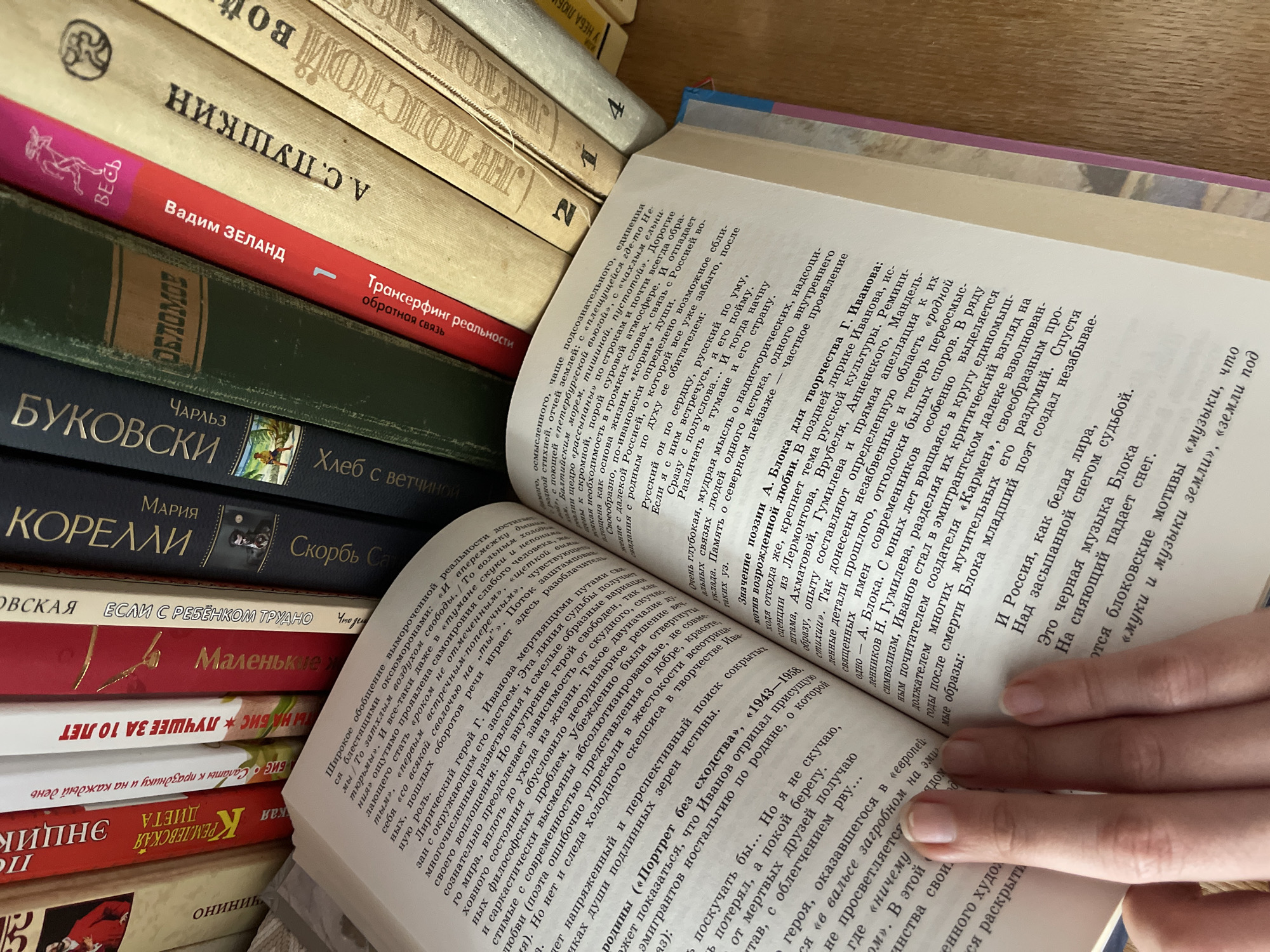 The image shows a close-up, overhead view of an open book with its pages fanned out. The book's pages are filled with dense Russian text. To the right and behind the open book, a stack of other books is visible, their spines facing forward and showing Russian titles and artwork. The spines are varied in color and design, with some appearing to be older than others. The lighting suggests an indoor environment, possibly a home or library. No specific location in the city of Sredny, Russia, can be determined from the image.