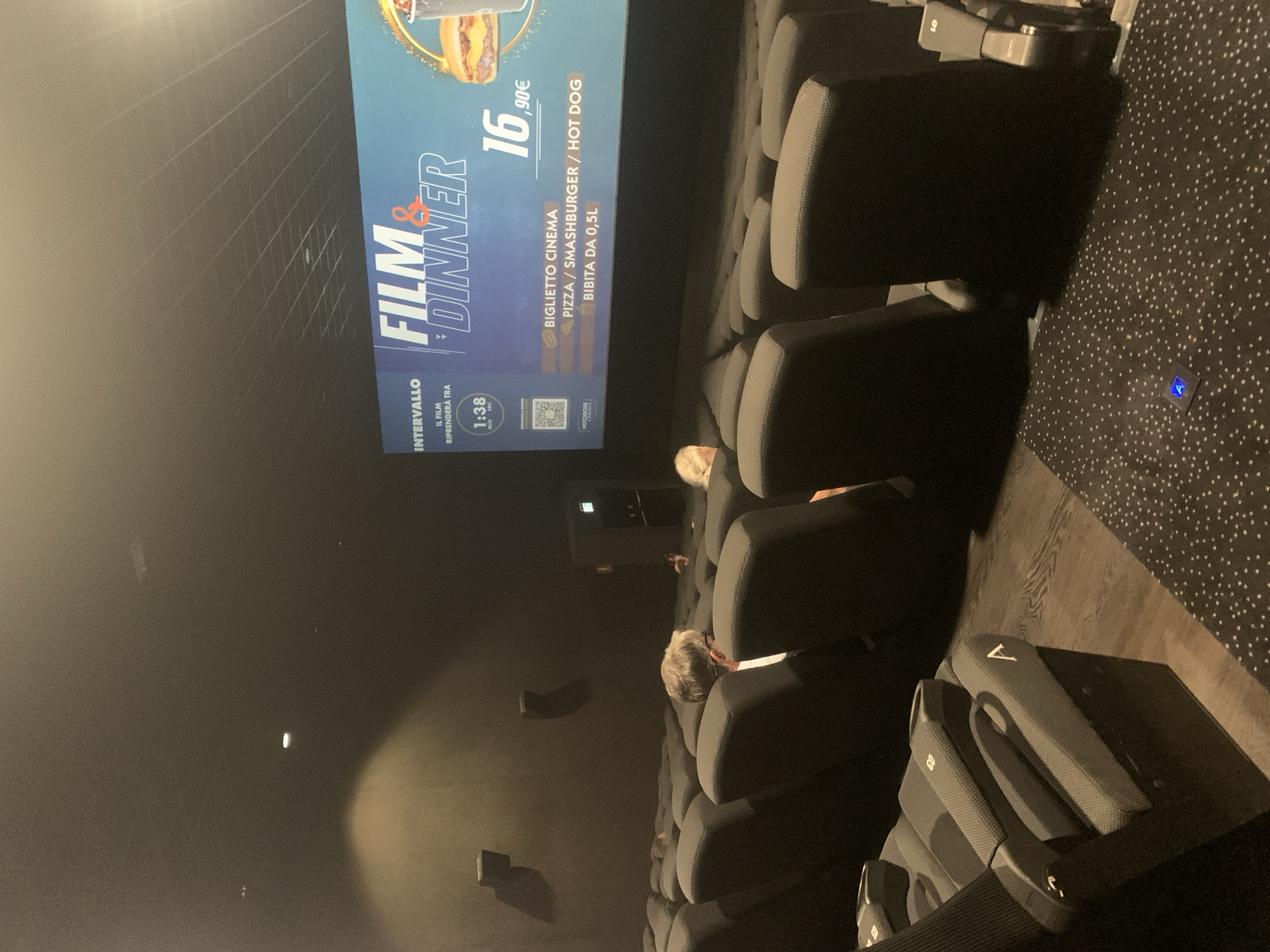 The image shows an interior view of a cinema auditorium in Sinalunga, Italy, during an intermission. The screen displays an advertisement for "Film & Dinner," indicating a movie screening combined with a meal. The advertisement features the price of 16.90€ for a cinema ticket that includes a pizza, smashburger, hot dog, and a 0.5L drink. A timer on the screen shows "1:38" for the remaining intermission time. Several rows of empty, dark-colored cinema seats are visible in the foreground and mid-ground. Two individuals are seated in the mid-ground, facing away from the camera, towards the screen. The environment appears dim, typical of a cinema setting. The floor in the foreground is carpeted with a dark pattern of white dots, and in the background, it appears to be wooden flooring.