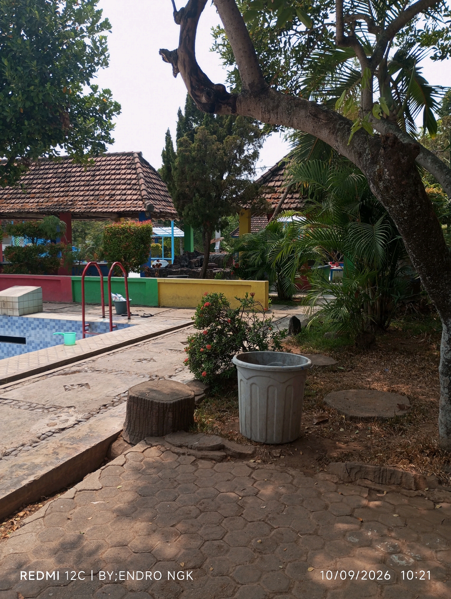 The image shows an outdoor scene in what appears to be a residential or recreational area in Sombron, Indonesia. In the foreground, a paved pathway with hexagonal tiles leads towards a swimming pool. Adjacent to the pool is a red ladder. A large, light gray cylindrical container sits on the paving stones to the right, with a small bush bearing red flowers next to it. Further into the background, there are several buildings with red-tiled roofs, surrounded by lush greenery and trees. The swimming pool itself has blue tiles and a concrete border. A white rectangular object is visible near the pool's edge on the left. The overall atmosphere is sunny and relaxed, suggesting a warm climate.