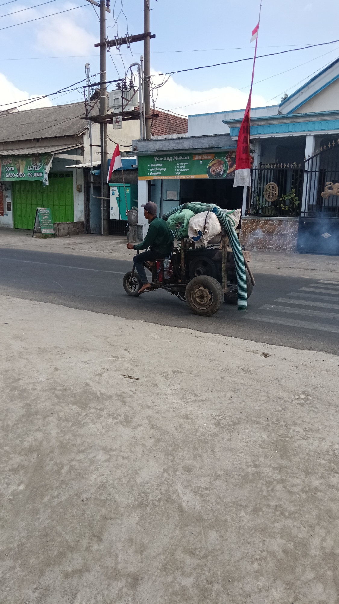 In Branggahan, Indonesia, a person wearing a dark green long-sleeved shirt and a baseball cap is operating a three-wheeled vehicle on a paved road. The vehicle is loaded with several large sacks and large flexible pipes. The person is riding on a small seat towards the front of the vehicle, with their feet resting on the ground. Behind the person, a white flag with red stripes and a red flag with white stripes are attached to poles. To the right of the road, a building with blue trim and decorative metal fencing is visible. In the background, there are other buildings and utility poles with electrical wires. Smoke is emanating from the right side of the image. A pedestrian crossing is marked on the road in the foreground.