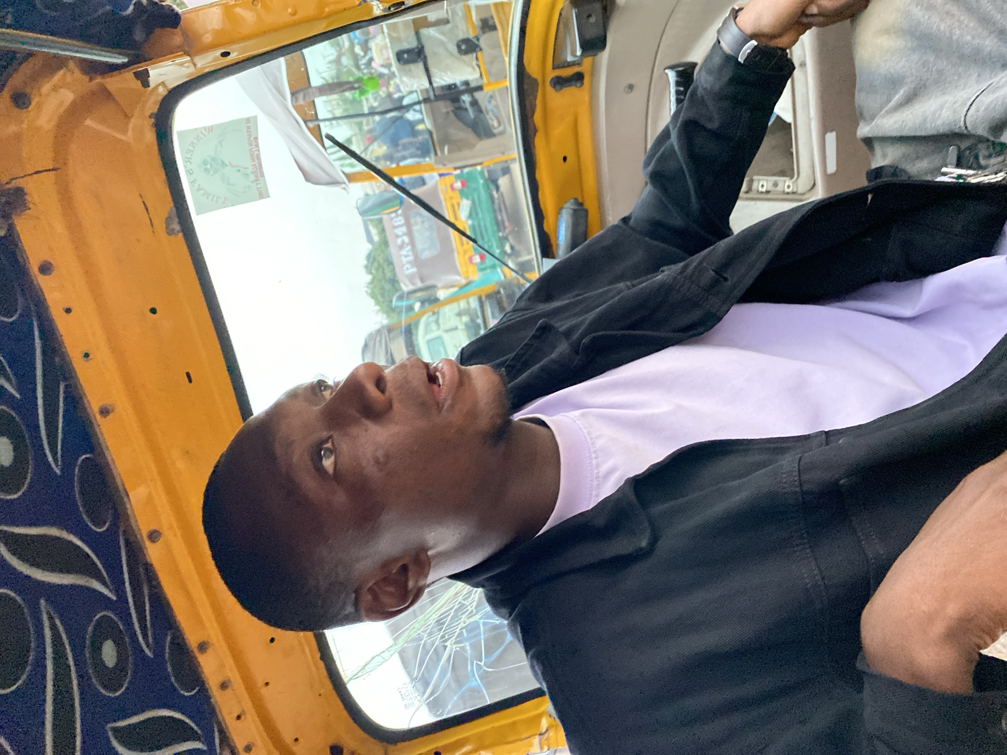 A young Black man, appearing to be in his early twenties, sits in the driver's seat of a yellow auto-rickshaw, likely in Bukuru, Nigeria. He wears a black jacket over a light purple t-shirt and a dark grey garment, possibly pants, is visible. His mouth is slightly open as he looks to his right, suggesting he is speaking or observing something outside the vehicle. The interior of the auto-rickshaw is yellow with a decorative blue and black patterned ceiling. The windshield is slightly cracked, and a sticker with an image and text is affixed to the upper right portion of the windshield. Through the windshield, other auto-rickshaws and people are visible, indicating a busy, urban environment. The sky appears overcast.