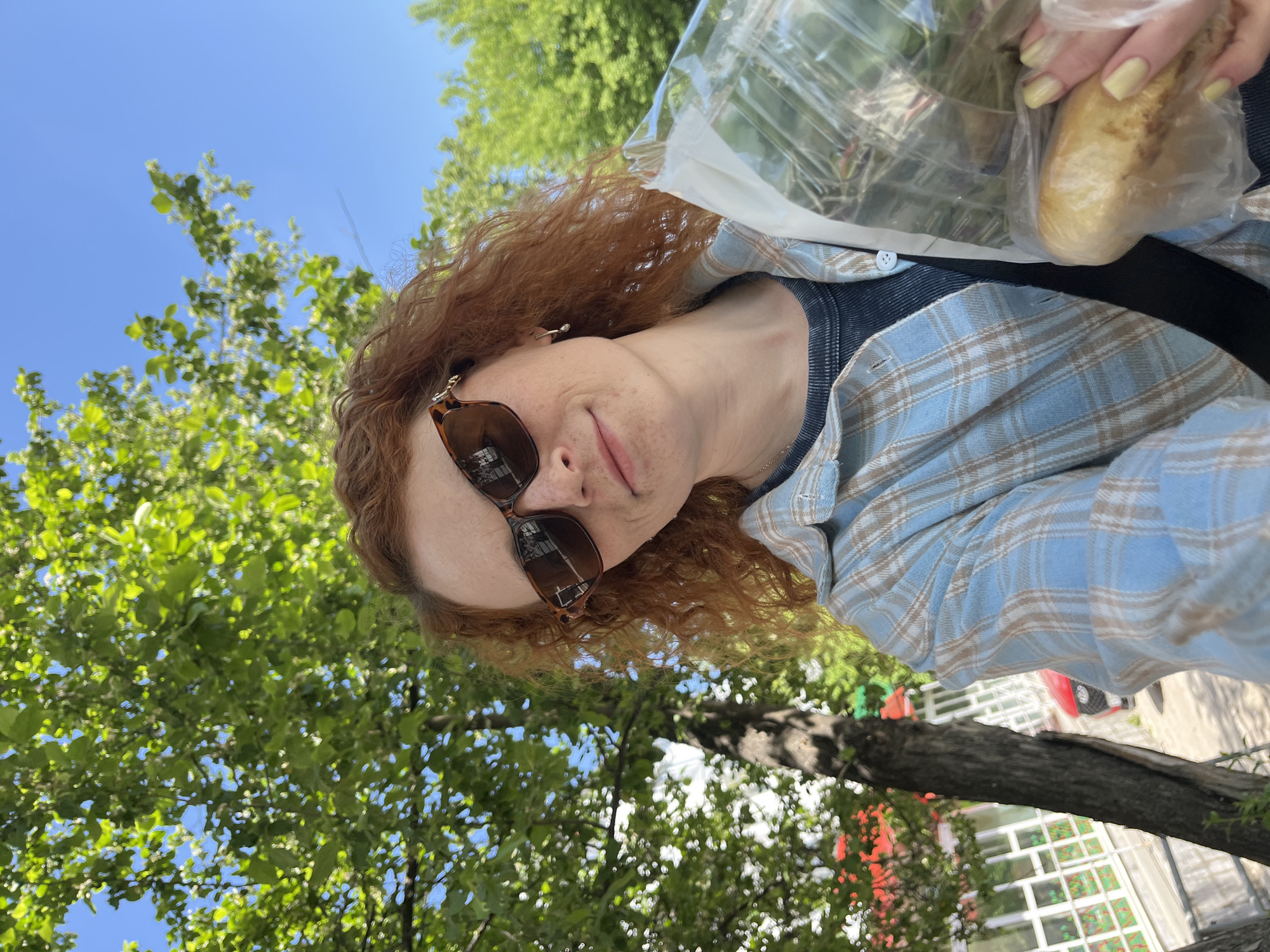 A person with red curly hair, wearing sunglasses and a blue plaid shirt, is holding a packaged loaf of bread. The person is standing outside, with trees and a blue sky visible in the background. A glimpse of a building structure with white frames and red accents is visible. The image appears to be taken in Chaikovsky, Russia, based on contextual information.