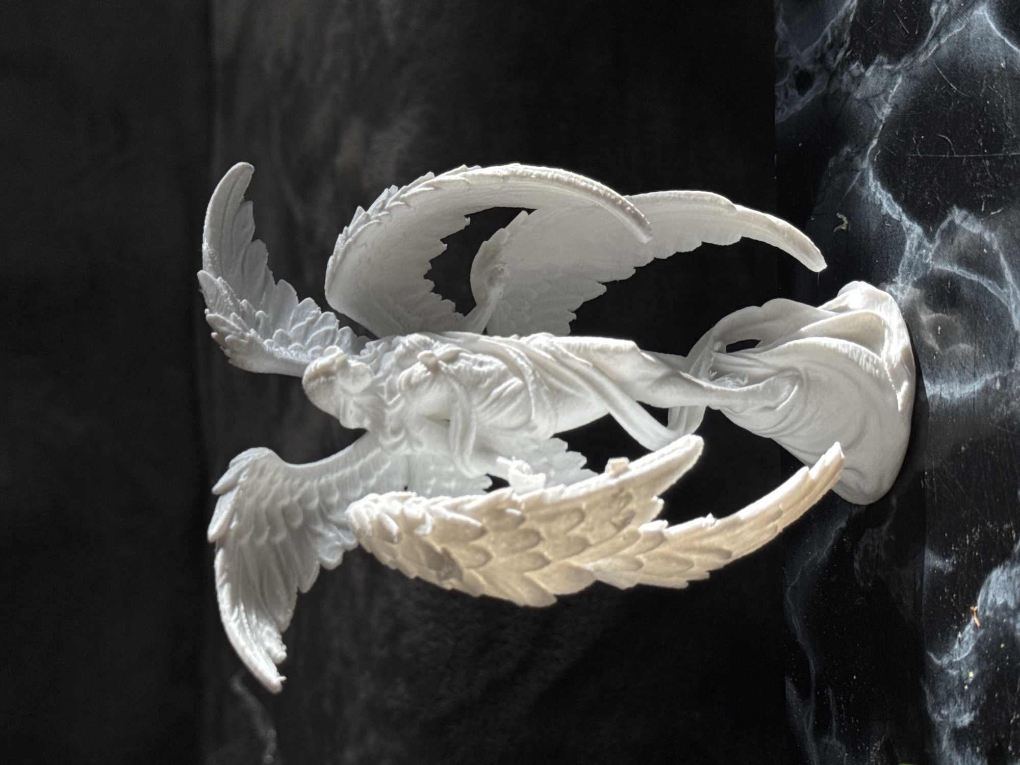 The image shows a white angel statue attached to a dark marble monument. The statue has four wings and appears to be in motion. The statue is located in the city of Sofia, Bulgaria, as indicated by its style and architectural context. The environment is dimly lit, possibly within a cemetery or memorial park. There are no other people or subjects in the frame. The focus is solely on the statue and its immediate surroundings.