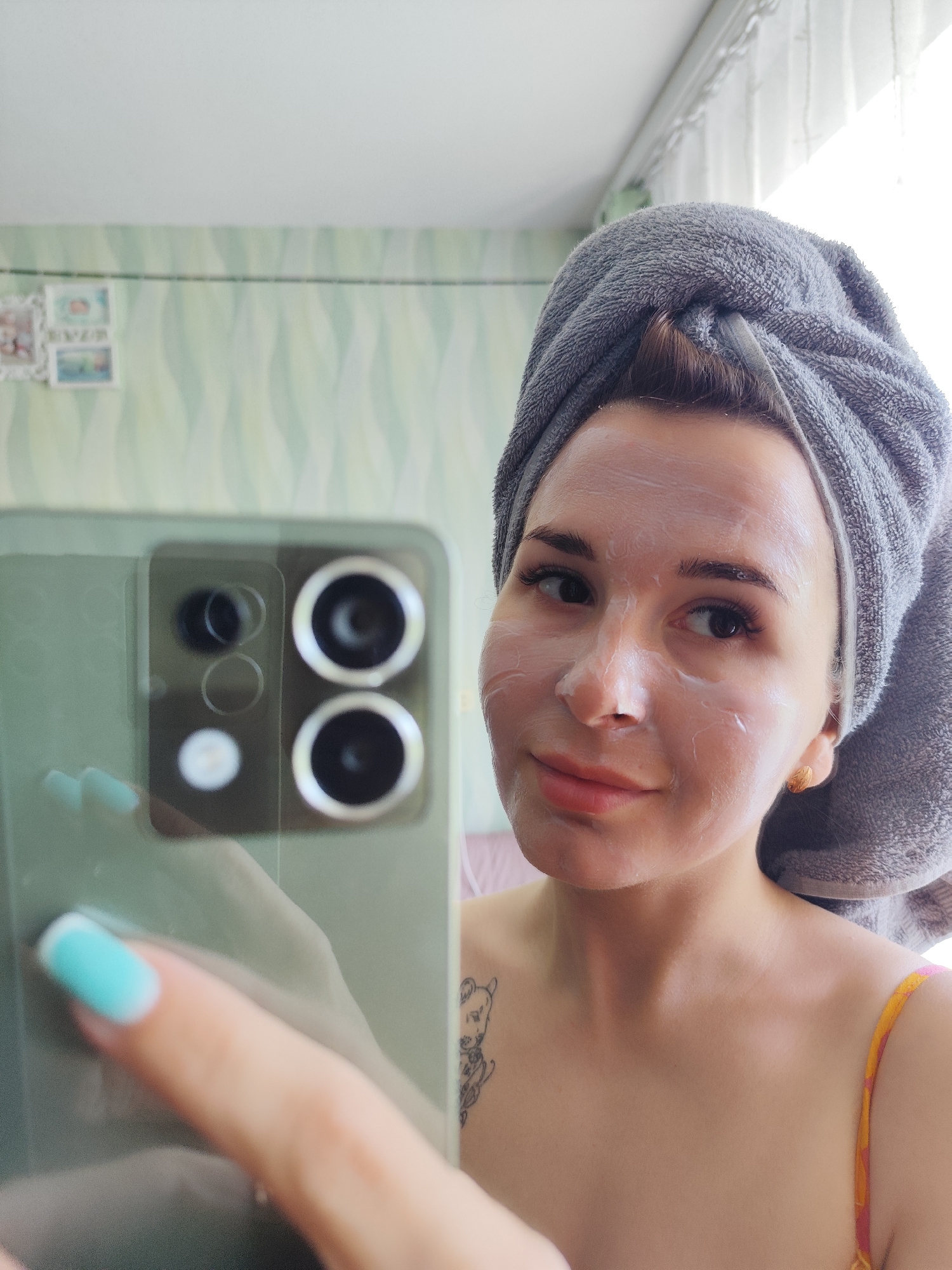 A woman wearing a gray towel wrapped around her head and a light-colored face mask is taking a selfie with a cell phone. Her nails are painted with light blue and white polish. She has a tattoo on her upper chest. The background appears to be an interior setting with patterned wallpaper and curtains. It is impossible to determine the location in the city of Фоки, Russia from this image.
