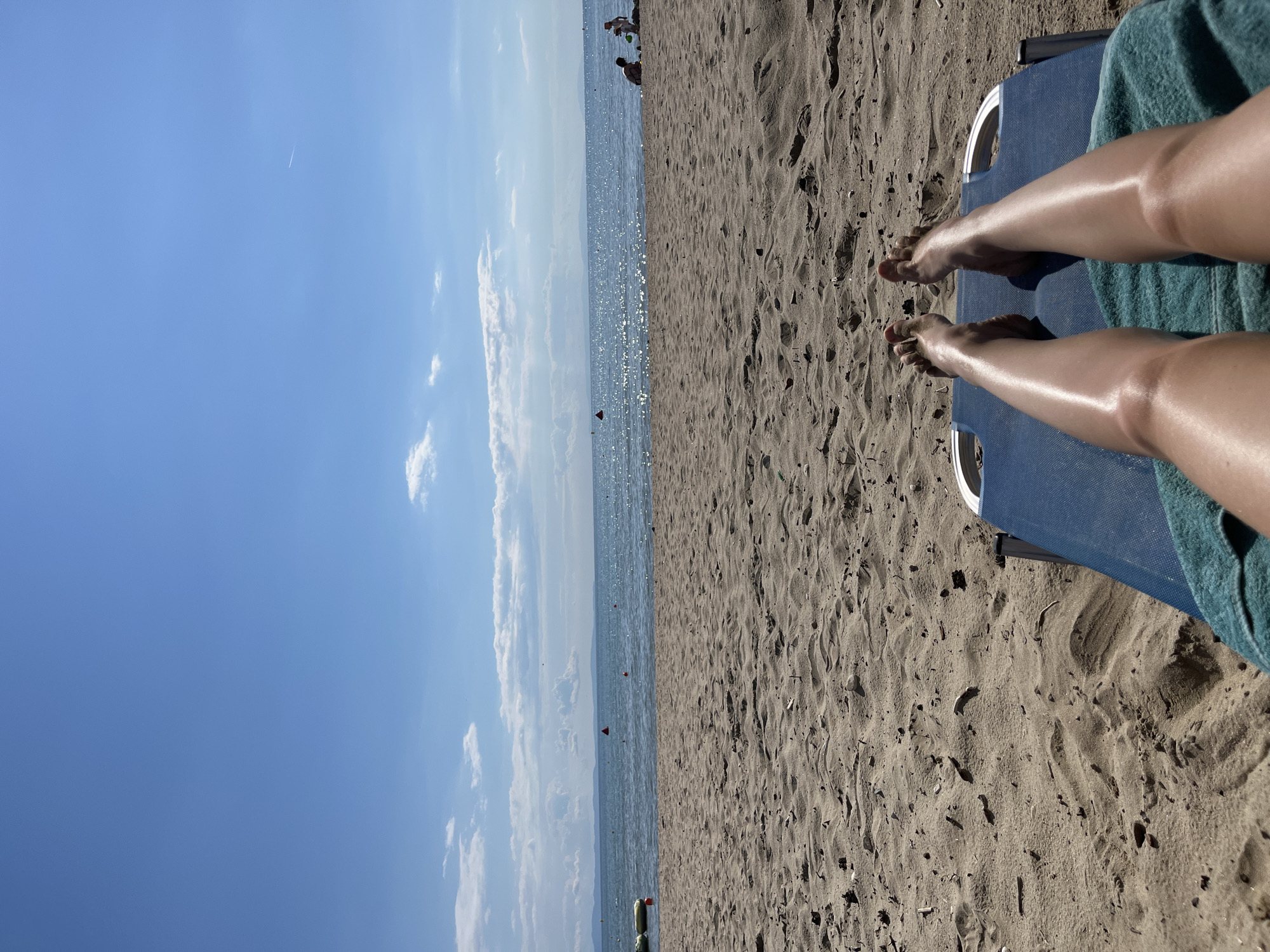 An overhead shot captures a pair of bare legs and feet resting on a blue lounge chair covered with a teal towel. The legs are tanned and appear moisturized or oiled, with the sunlight reflecting off them. The lounge chair is positioned on a sandy beach. The sand is light brown and appears somewhat rippled, with scattered debris like small sticks and leaves. Beyond the beach, a calm, blue body of water stretches towards a hazy horizon. The water has shimmering reflections from the sun. Several small red buoys are visible in the water. In the distance, on the right side of the frame, two figures are seated on the sand near the water's edge, and another person is standing. The sky is a clear, bright blue with a few scattered white clouds. A faint contrail is visible high in the sky on the right. The overall scene depicts a sunny day at the beach. The location is identified as a beach in an unspecified part of Greece.