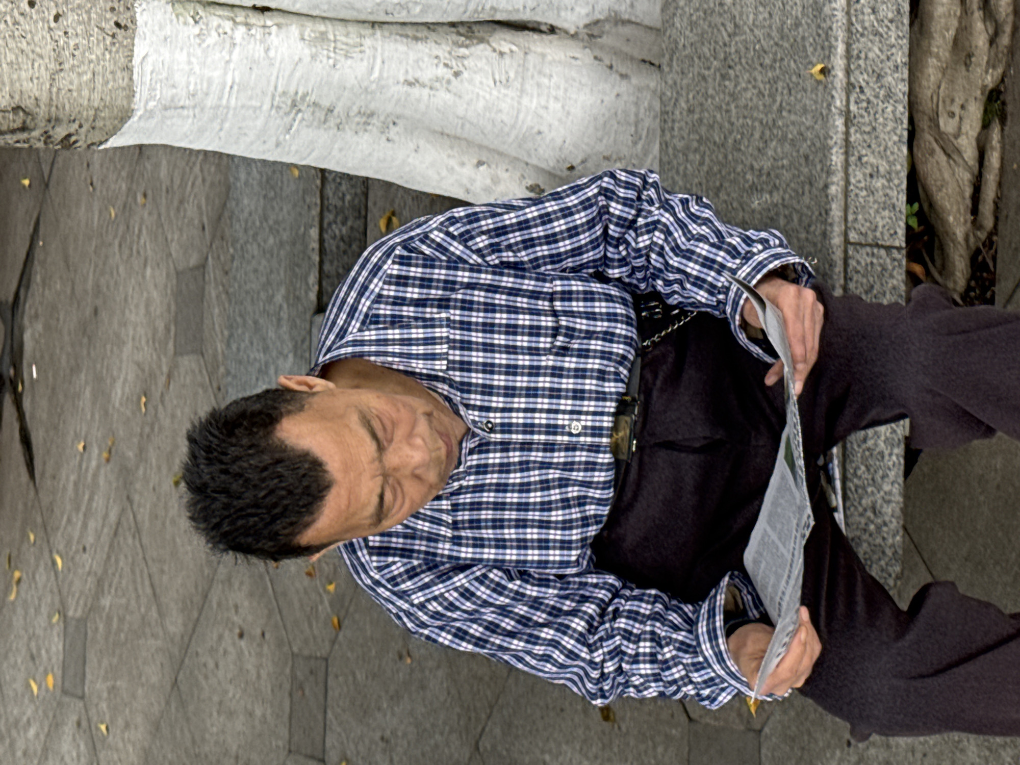A man is seated on a stone structure of Jiangjin, China, intently reading a newspaper. He is wearing a blue and white checkered long-sleeved shirt, dark-colored pants, and has short, dark hair. He sits next to a concrete structure with white paint on it and an older stone edge. The surrounding area is paved with stone blocks. A tree trunk and a few small plants are visible nearby.