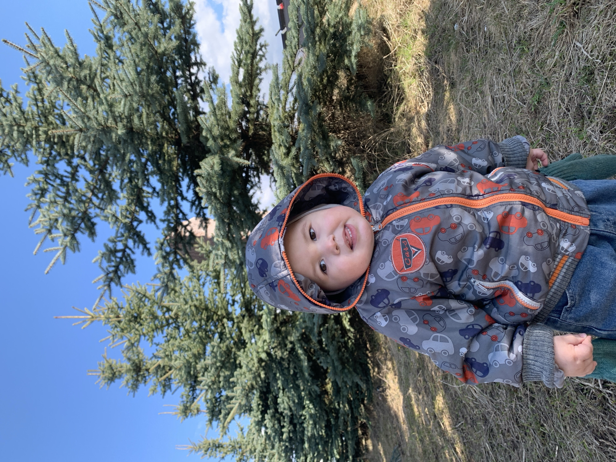 A young child in a car-patterned jacket and hood is lying on the ground near a pine tree, smiling at the camera. The child is wearing blue jeans. The background consists of pine trees and a blue sky with scattered clouds. The ground has patches of dry grass.
It is not possible to determine the location is in the city of Фоки, Russia from the image.