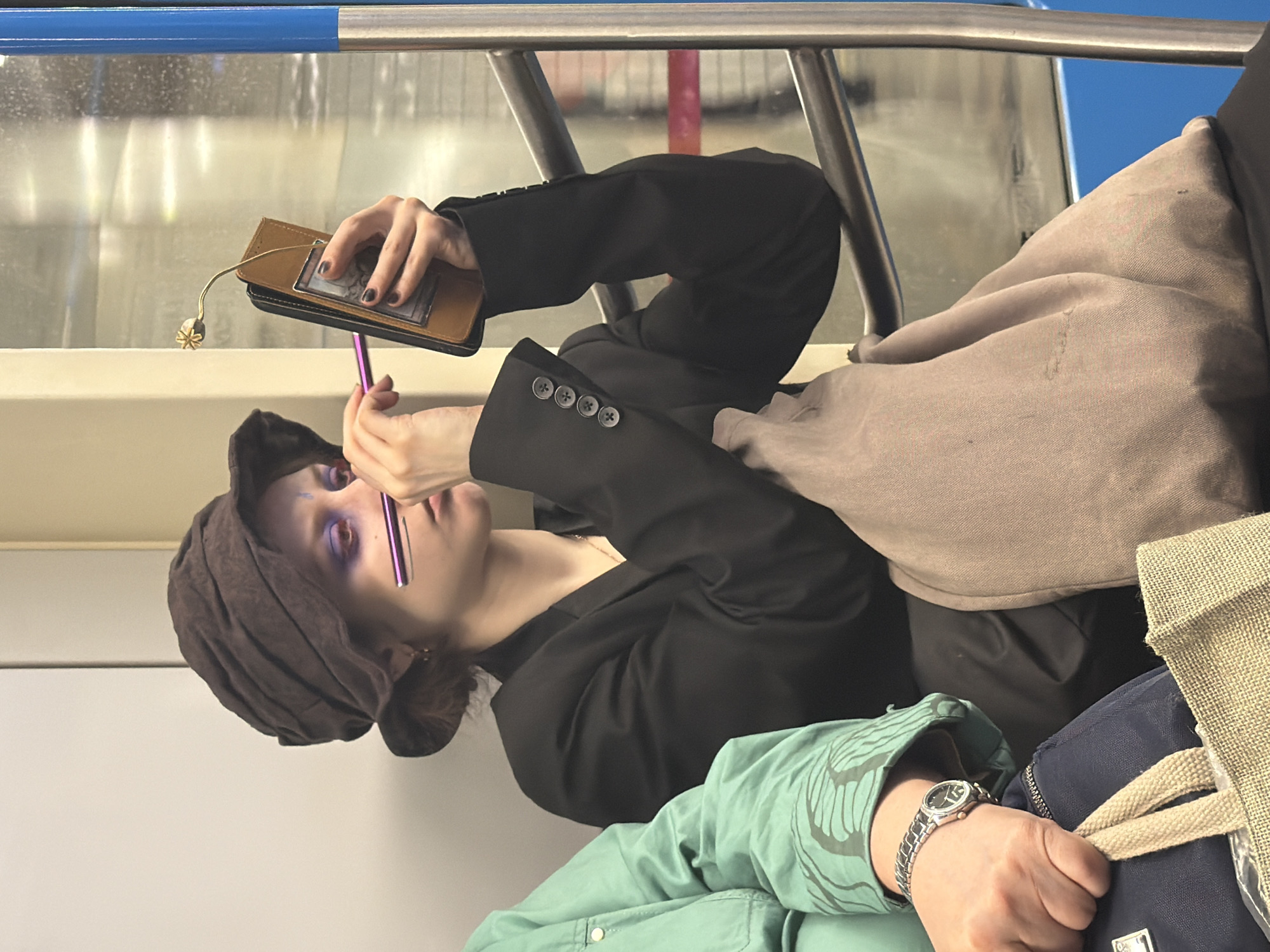An image captures a moment inside what appears to be a public transport vehicle, likely a train or subway car, given the blue and metallic handrails and window reflections. The scene is set in Moscow, Russia.
The primary subject is a person seated, seen from the chest up, engaged in applying makeup. They wear a dark, possibly brown, head covering that wraps around their head and a black jacket with buttons visible on the sleeve cuff. Their eyes are strikingly made up with vibrant purple or blue eyeshadow. In their left hand, they hold a brown and black wallet-like object with a small, light-colored charm resembling a poppy seed pod dangling from it. Their right hand holds a pink or purple makeup pen, which they are using to carefully apply makeup to their eye.
In the bottom right foreground, the arm of another person is visible. This individual wears a teal or light green jacket with a faint, light-colored pattern on the sleeve. A silver-toned wrist watch with a dark face is worn on their left wrist. Their hand grips the beige strap of a navy blue bag.
The background shows reflections in the vehicle's window and the interior structure, including a light-colored wall or partition and the metallic support bars of the seating area. The overall impression is that of a commuter utilizing travel time for personal grooming. There is no visible text.