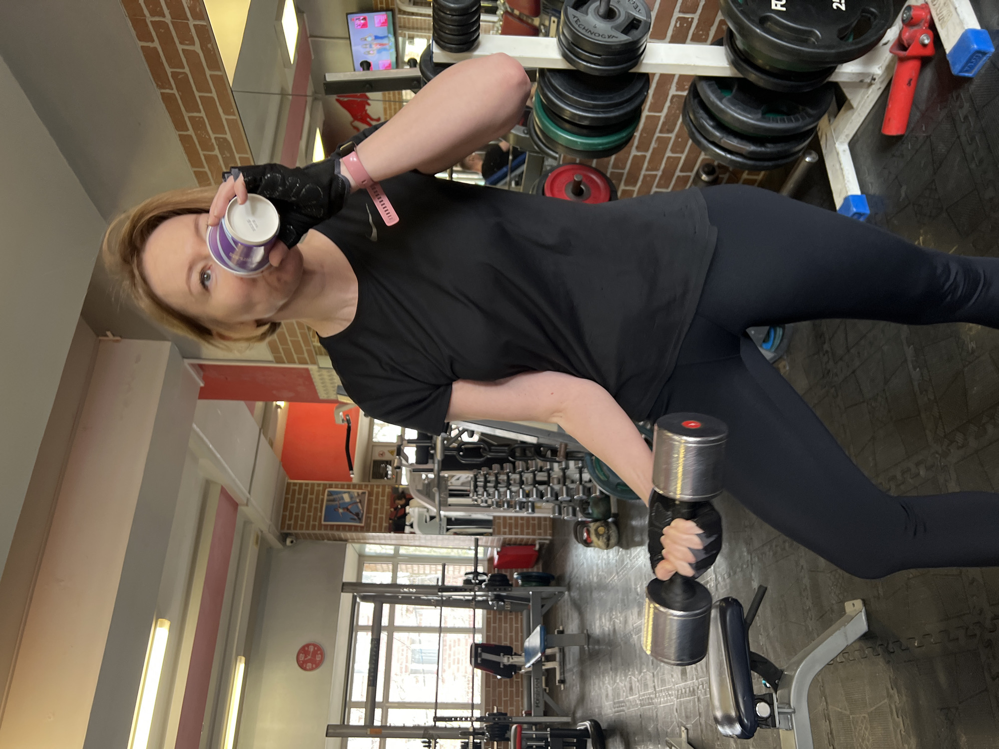 The image shows a woman in a gym, likely in Saint Petersburg, Russia, pausing during or after a workout. She is standing, looking towards the viewer, and is dressed in black athletic attire including a short-sleeved top with a faint white logo on the left chest, black leggings, and black fingerless workout gloves. A pink fitness tracker is visible on her left wrist.
In her right hand, she holds a silver metal dumbbell, with her arm resting on a piece of gym equipment. With her left hand, she holds a small, white cup with purple branding to her mouth, as if drinking from it.
The gym environment features various pieces of exercise equipment. Immediately behind the woman is a rack stacked with black weight plates, some of which are clearly marked with "TECHNOGYM." The wall behind this rack has a distinct red brick pattern. The floor is covered with black interlocking foam mats. In the background, large windows allow natural light into the space, and a red circular clock is visible on a far wall. Other gym machines and benches can be seen in the distance, alongside a further brick-patterned wall section. The lighting suggests it is daytime.