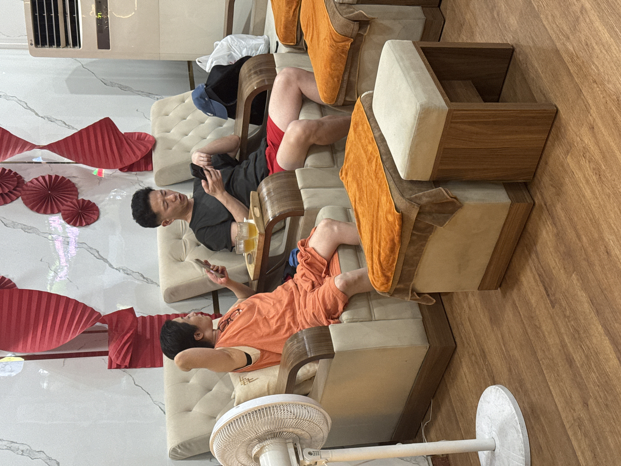 Two individuals are seated in comfortable armchairs within a spa-like environment in Nha Trang, Vietnam. On the left, a woman wearing an orange t-shirt and shorts is holding a mobile phone in her left hand, with her arm raised behind her head. On the right, a man in a black t-shirt and red shorts is also looking at his mobile phone. A tray with two glasses of amber liquid is placed on the armrest between them. In the foreground, there are footrests covered with orange towels and a small wooden stool with a cream-colored cushion. A white oscillating fan stands to the left of the woman. The walls are decorated with red paper fan-shaped ornaments and feature a marble-like pattern. An air conditioning unit is visible on the far right. The floor is made of polished wood.