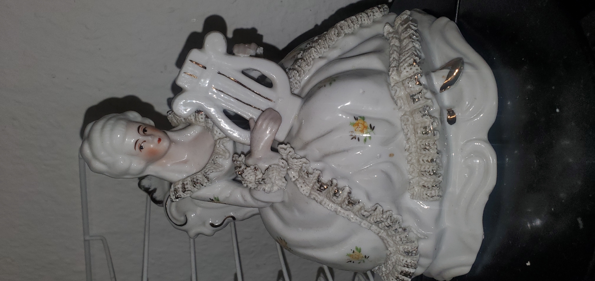 A white porcelain figurine of a woman with a powdered wig and an ornate dress sits holding a lyre. The woman's eyes are looking forward, and her lips are painted red. The dress has ruffled trim with gold accents. Small yellow flowers are painted on the dress. The base of the figurine is white and wavy, with gold accents on the edge. The figurine is on a dark, reflective surface. The background is a light-colored wall with some shadows.