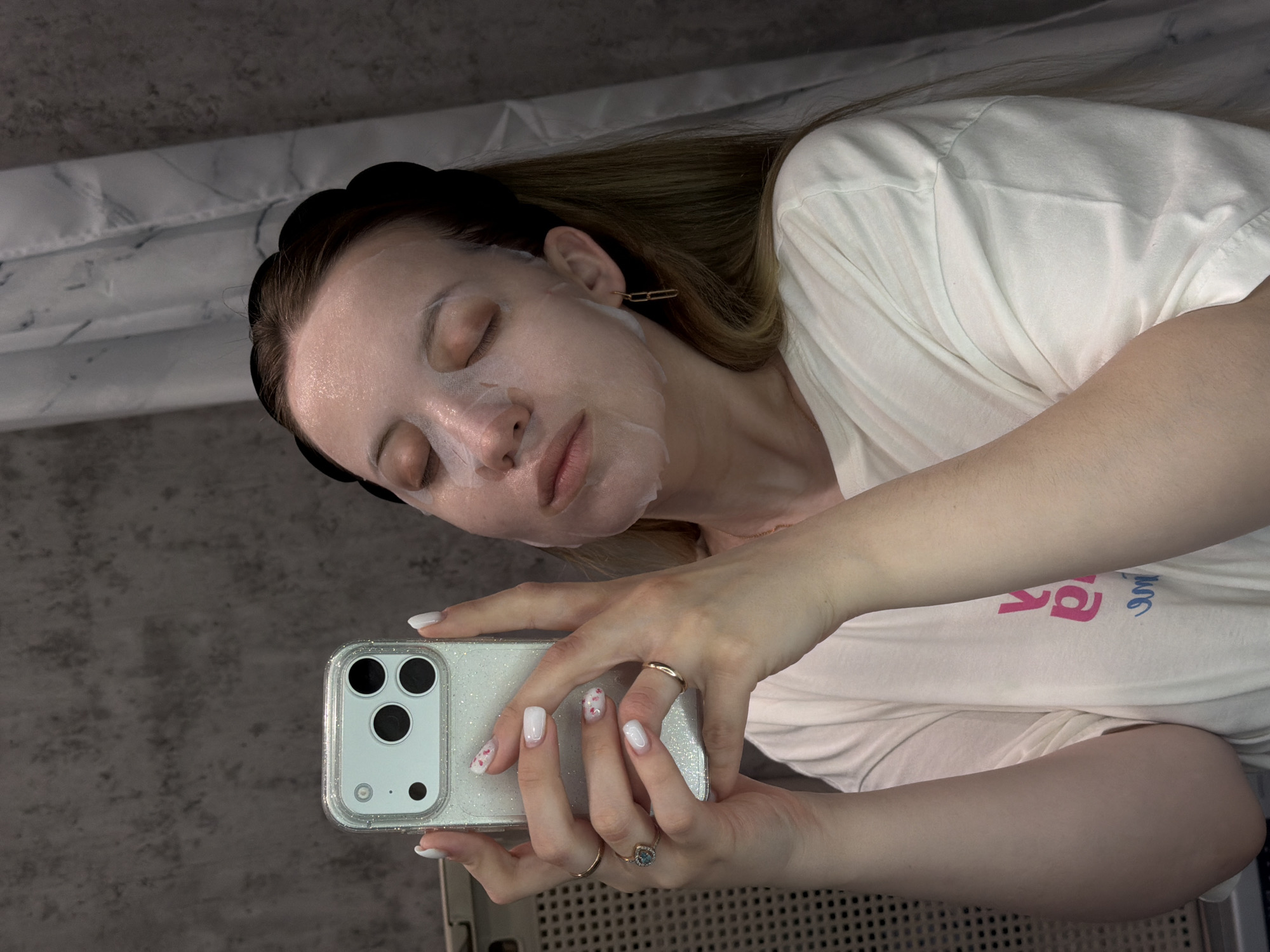 A woman with a sheet face mask and a black headband is taking a selfie. She is holding a white smartphone with a clear, glittery case. She has white nail polish with pink floral designs on some nails, and she wears two rings, one gold and one with a blue stone. She is wearing a white t-shirt. The background appears to be a bathroom with a gray wall and a marble-patterned surface. The location is Reshetnikovo, Russia, although this information is impossible to determine from the image alone.
