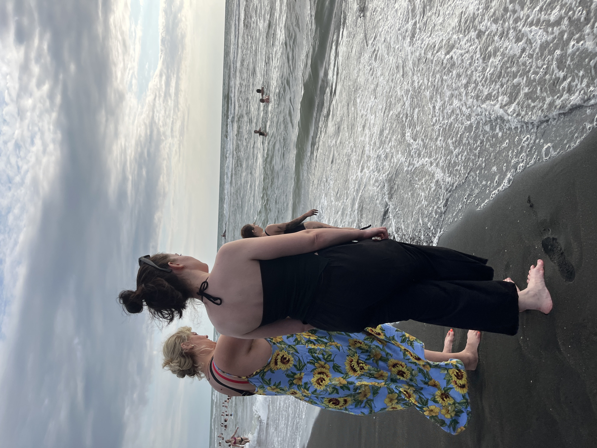In Ureki, Georgia, two women stand on a dark sandy beach, their backs to the camera, looking out at the sea. The woman on the left wears a blue dress with a sunflower pattern and a red-and-white striped bikini top. The woman on the right is dressed in a black strapless top and black pants. The sea is choppy with moderate waves, and several people are visible swimming in the distance. The sky is overcast with soft, diffused light. The scene captures a moment of leisure on a beach, with the natural environment of the sea and sky playing a significant role.