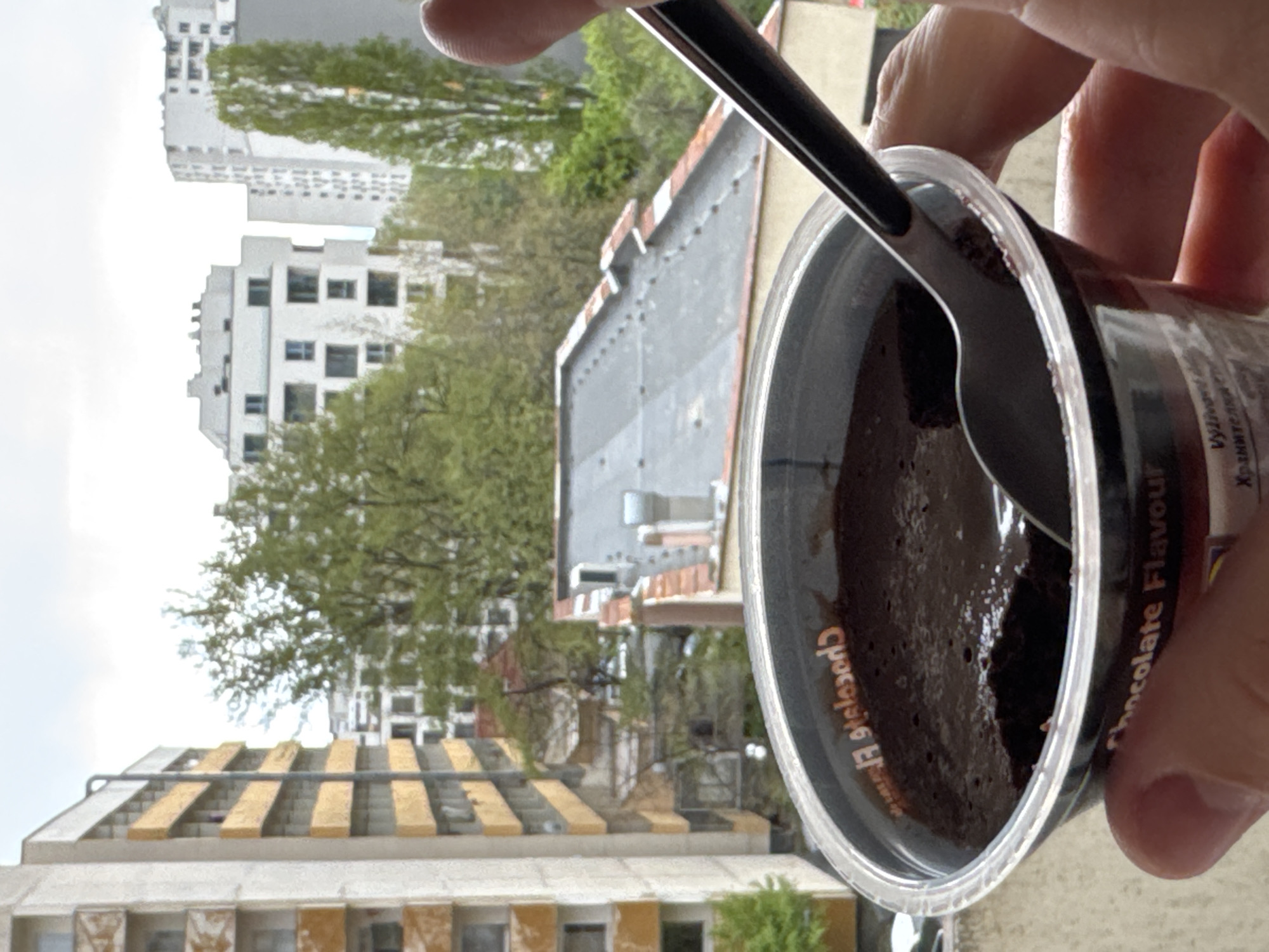 A hand holds a cup of chocolate dessert with a spoon inside, with a view of Sofia, Bulgaria in the background. In the immediate background, a building with a gray roof and a row of trees. In the far background, buildings with a light facade. The image is taken during the day, and the lighting appears to be overcast. The dessert appears to be a chocolate mousse. The writing on the cup includes the phrase "Chocolate Flavor". The buildings in the background suggest an urban setting. The perspective is at eye-level, looking out from an elevated position such as a balcony or high window. The urban environment suggests the buildings are located in Sofia.