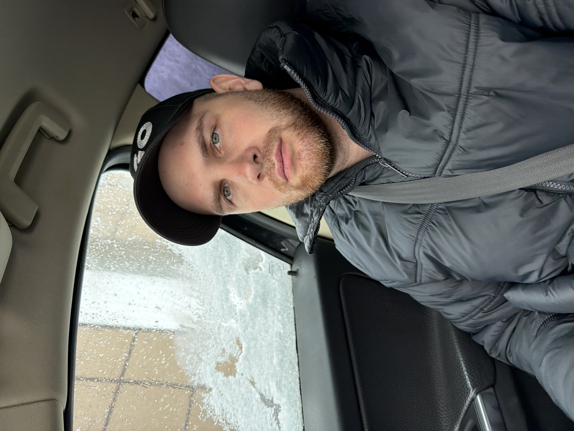 The image is a selfie taken from inside a car. A man with a red beard and wearing a black baseball cap and a gray puffer jacket is visible. He is wearing a seatbelt. The car window shows rain or melting snow. Based on the location provided, the photo appears to be taken in Chaikovsky, Russia.