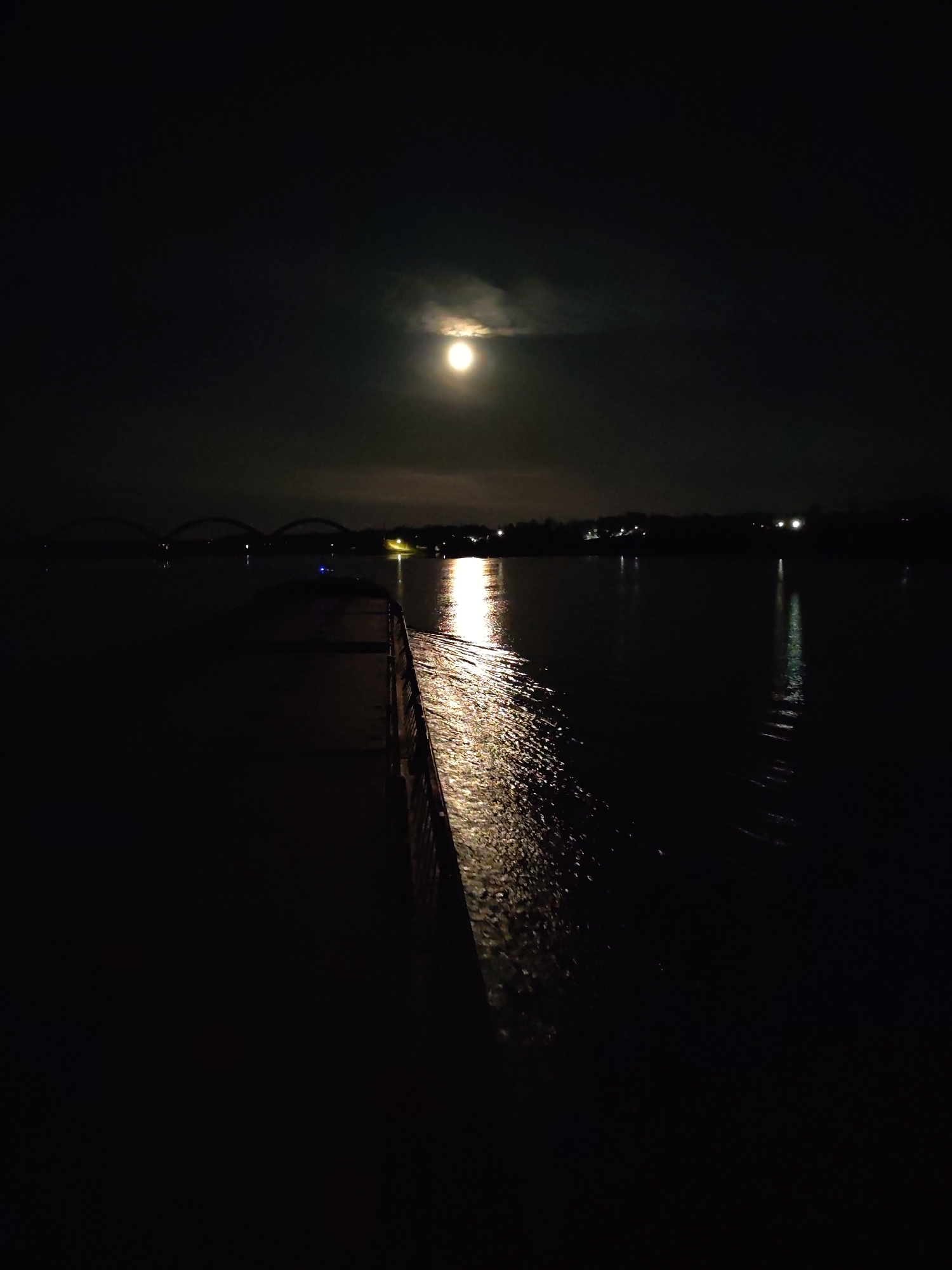 A night-time photograph shows a body of water, likely a river or lake, with a pier or dock extending into the water. The moon is visible in the sky, partially obscured by clouds. The reflection of the moon is visible on the water's surface. There is a bridge visible in the distance with multiple arches. Across the water, there are lights indicating a town or city. Based on the location, the picture was likely taken near or on the river or lake, somewhere in or near Gladyshevo, Russia. There are no people visible. The environment is dark and the scene appears quiet.