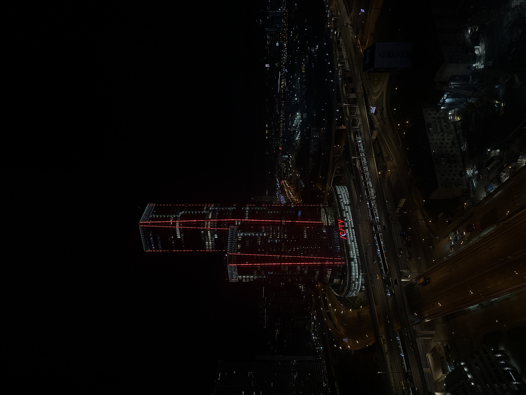 This high-angle, nighttime photograph captures a dense urban landscape in Khimki, Russia. The scene is dominated by artificial light sources against a deep black sky, characteristic of a clear night.
In the mid-ground, a complex network of multi-lane highways and overpasses stretches across the frame, illuminated by numerous streetlights and the moving lights of vehicles, which appear as streaks and dots. These roads suggest a significant transportation hub.
Two prominent, modern high-rise buildings stand out towards the left. Both are adorned with distinctive horizontal red illuminated lines across their facades, adding a striking visual element to the night skyline. On one of these central buildings, the text "/CITY" is clearly visible in large, red, illuminated letters. Other, less defined buildings with illuminated windows are scattered across the urban sprawl, extending into the background, showcasing the city's vastness.
The overall impression is one of a bustling and modern city, active even late at night, with its infrastructure brightly lit, showcasing its scale and contemporary architecture under a clear night sky.