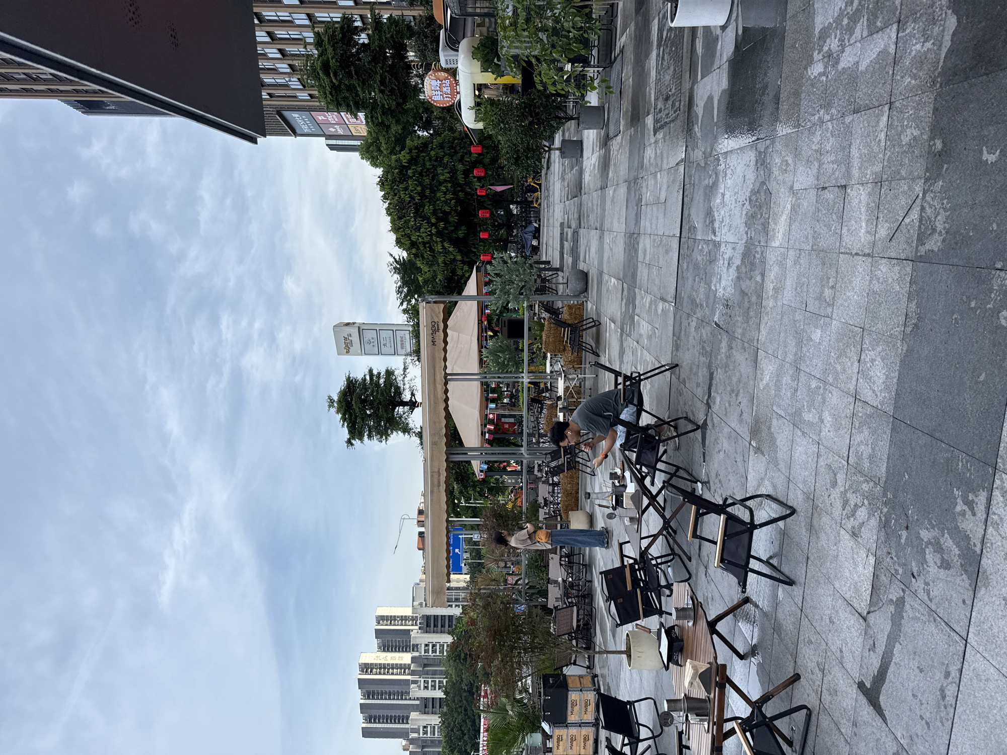 This image captures an outdoor seating area of a restaurant or cafe, likely in Haizhu District, China, with a contemporary urban backdrop. The scene is set on a paved stone walkway, with some areas showing signs of recent rain, creating a wet, reflective sheen.
The primary subjects are two individuals preparing the area. One person, wearing a grey t-shirt and shorts, is seen interacting with a table and chairs, possibly arranging them for service. Another person, a woman in a light-colored top and blue jeans, stands nearby, also looking towards the tables. The seating area is furnished with numerous black metal chairs and wooden tables, some stacked or folded, indicating it might be early morning or that preparations are underway. A few straw-filled ottomans are also visible.
In the background, a building with glass windows and signage suggests a commercial district. Green trees provide a natural element, contrasting with the urban structures. Red lanterns are strung across an awning, adding a touch of festive decoration. The sky is partially cloudy, with a soft, diffused light, suggesting it could be morning or late afternoon.
Visible text includes "MWBBQ" printed on several wooden crates, likely related to the establishment. Another sign reads "TFRESH七鮮", identifying a supermarket chain. "太二" is also visible, potentially a restaurant name or part of a larger sign. The overall atmosphere appears calm and orderly, with a focus on setting up the outdoor space.