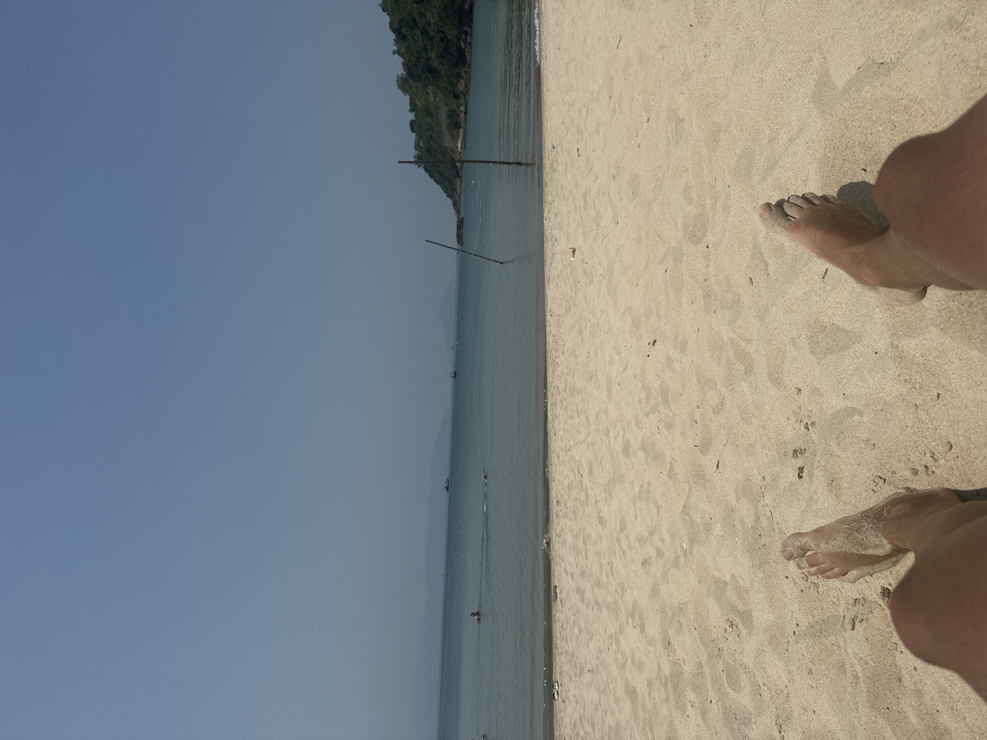 A first-person perspective image depicts a sunny beach scene on the Sơn Trà Peninsula in Đà Nẵng, Vietnam. The foreground features the bare, tanned legs and feet of an individual, partially embedded in light tan sand. The sandy beach transitions to calm, clear blue-green ocean water. Near the waterline on the sand, a black sandal and a white sandal are visible, along with a small white cylindrical object further to the right. In the midground ocean, a rudimentary volleyball net is partially submerged, held by two vertical wooden poles. Two individuals are present in the water: one is floating face-up with an inflatable ring, and another is swimming away from the shore. Several small boats are visible in the distant water. The background reveals a verdant, tree-covered hill rising from the ocean on the right side, while hazy distant mountains are visible across the bay on the left horizon. The sky is clear and light blue.
