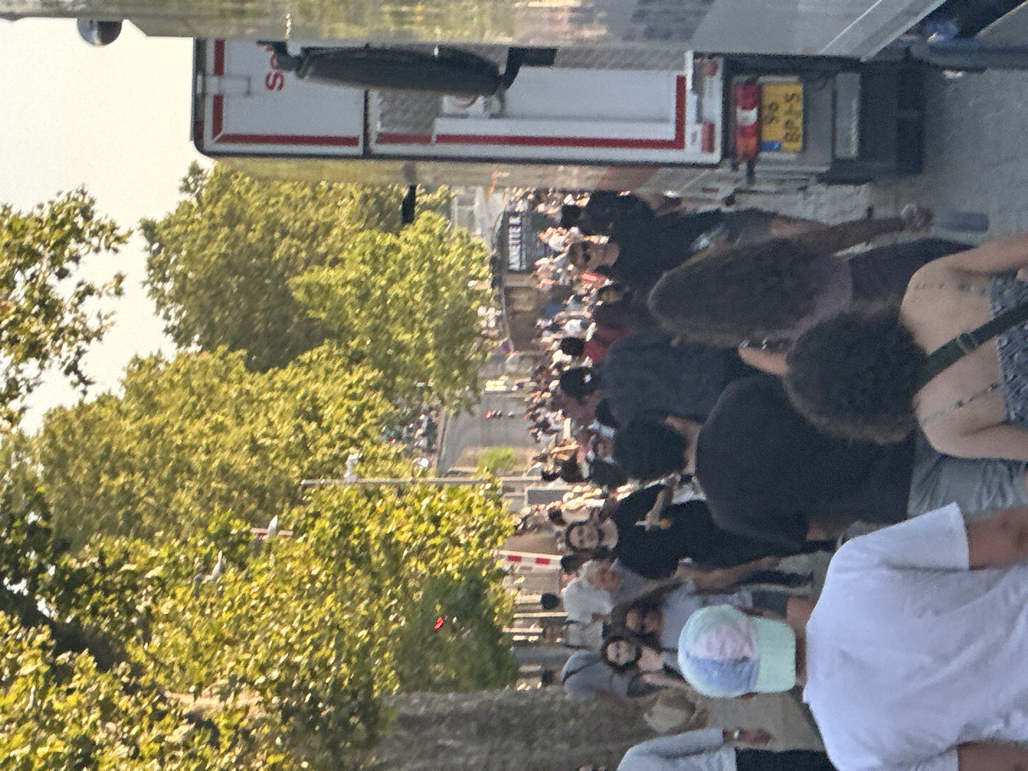 A street scene in Paris, France, shows a crowd of people walking on a sidewalk lined with trees. A large white truck with red trim is parked on the right side of the image, its side visible. The truck has a license plate that reads "86 BPJ 5". Many people are walking in both directions, appearing to be enjoying a sunny day outdoors. The trees are full of green leaves, and sunlight filters through them, casting dappled shadows on the ground. Some of the people are casually dressed, suggesting a relaxed atmosphere. The overall impression is of a lively public space during the day.