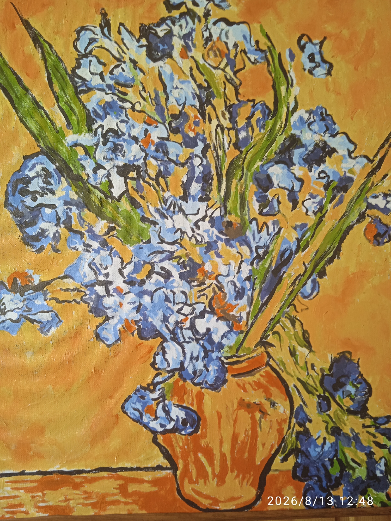 The image is a close-up, vertical, eye-level, medium shot of a painting of blue irises in a vase against an orange background. The painting style is impressionistic, with visible brushstrokes and bold outlines. The irises are rendered in various shades of blue, with some white and orange accents. Green leaves are interspersed among the flowers. The vase appears to be terracotta-colored and is partially obscured by the flowers. The background is a vibrant orange, with some lighter and darker variations suggesting texture. In the bottom right corner of the painting, a timestamp is visible, reading "2026/8/13 12:48". The painting appears to be on a surface with a visible wooden texture at the very bottom. There are no people, actions, or interactions depicted in the image. The location in the city of Chișinău, Moldova, is not discernible from the image itself.