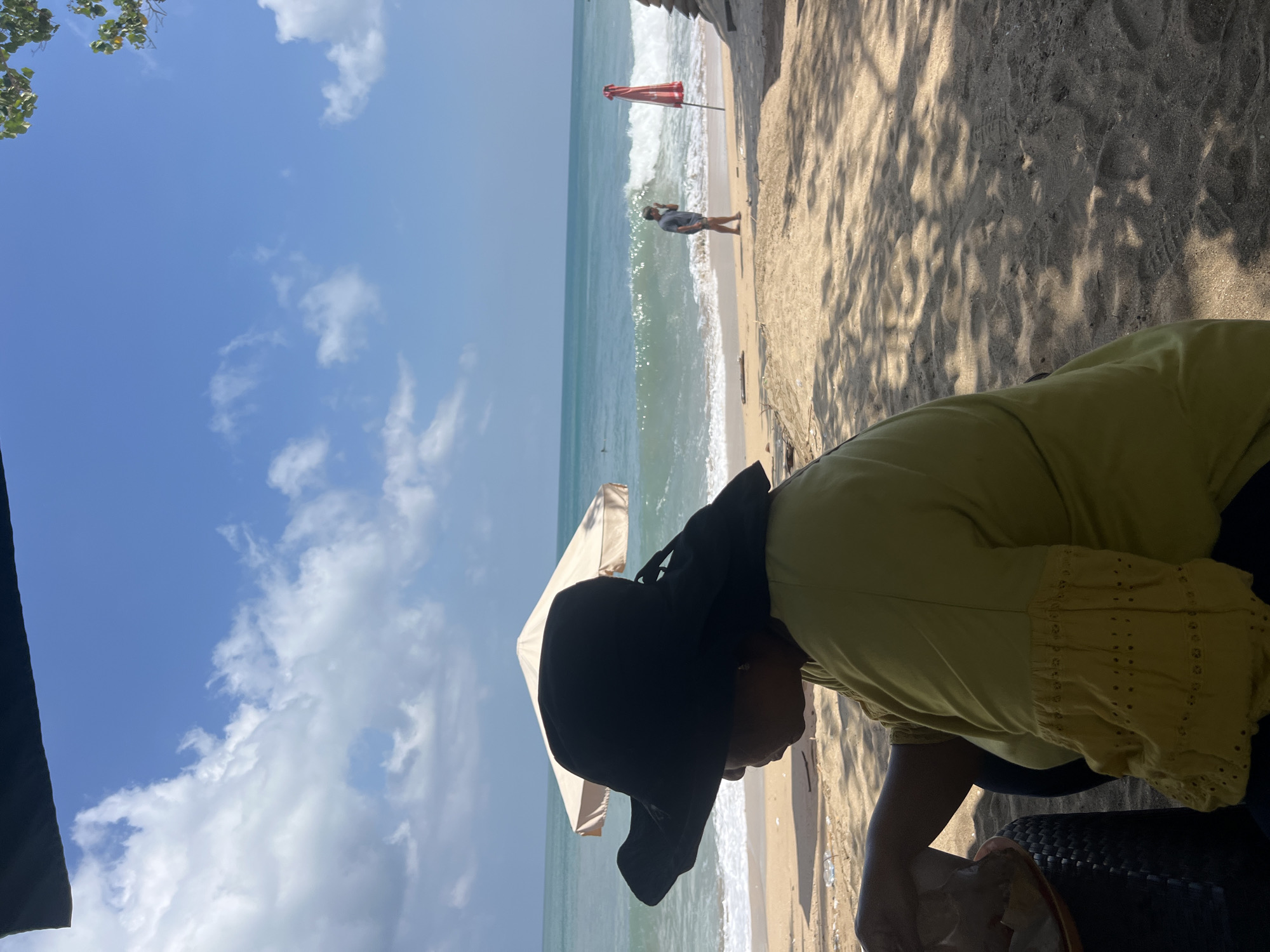 A woman wearing a wide-brimmed black hat and a yellow top sits in the foreground on a sandy beach. She is looking down and to her left, with her right arm resting on a dark woven chair and her left hand near a plate. In the background, the turquoise ocean meets a sandy shore, with gentle waves breaking. A beige beach umbrella stands to the left of the frame, and a red beach umbrella is visible further down the beach. A person in a grey t-shirt and shorts is walking near the water's edge. The sky is blue with scattered white clouds. The scene appears to be Kuta Beach, Bali, Indonesia, a popular tourist destination known for its surf and vibrant atmosphere.