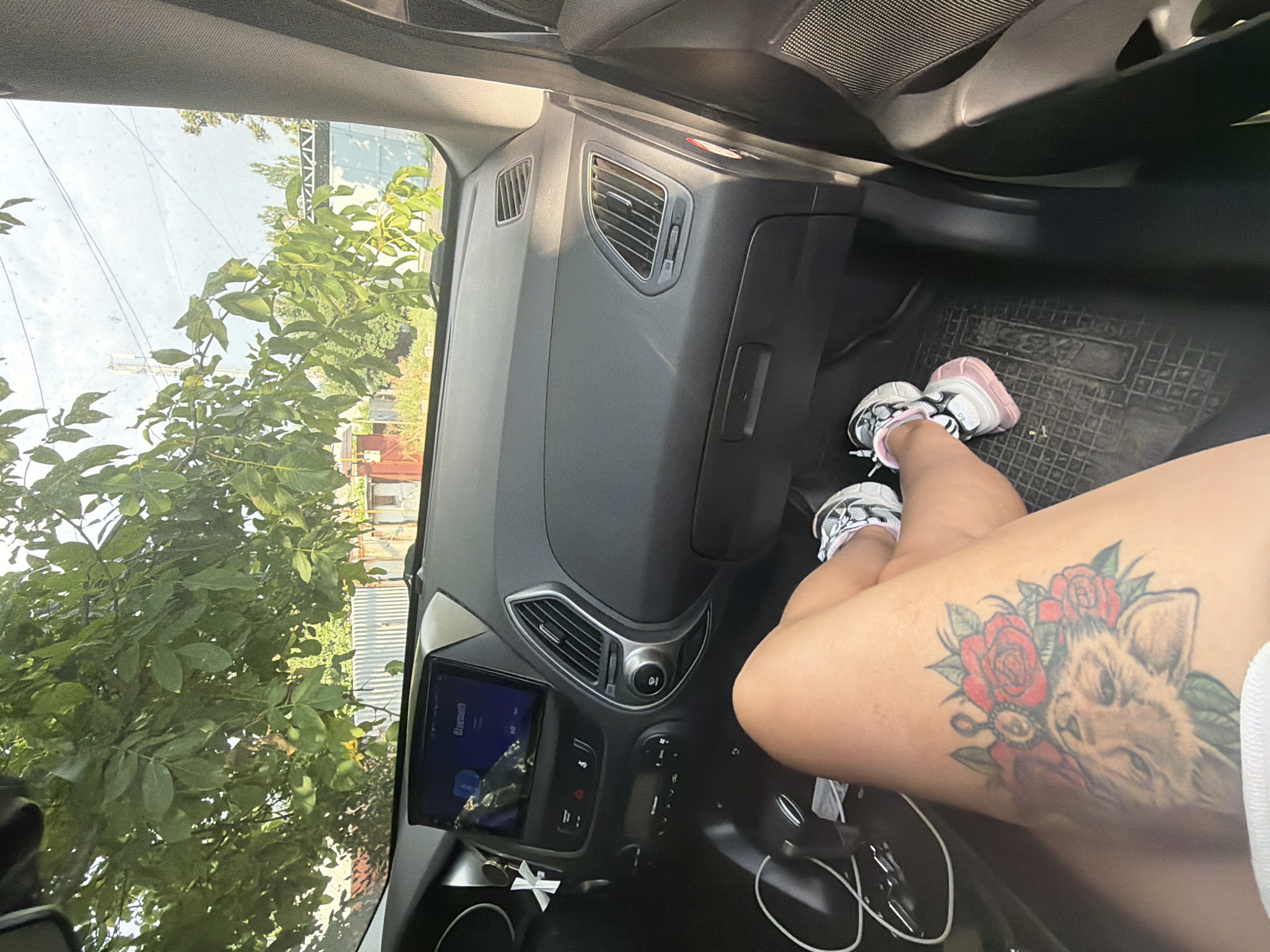 The image shows the interior of a car, looking out through the windshield. In the foreground, a person's legs are visible, resting on the passenger side of the car. They have a tattoo on their left thigh depicting a lion's head surrounded by roses. The person is wearing sneakers with pink accents. The car's dashboard is visible, including a touchscreen display. Outside the car, trees with green leaves hang over the windshield, and beyond that, there are buildings and a fence. The scene appears to be in a residential area, with a somewhat overgrown appearance. There is no specific indication that the location is in the city of Donetsk, Ukraine.