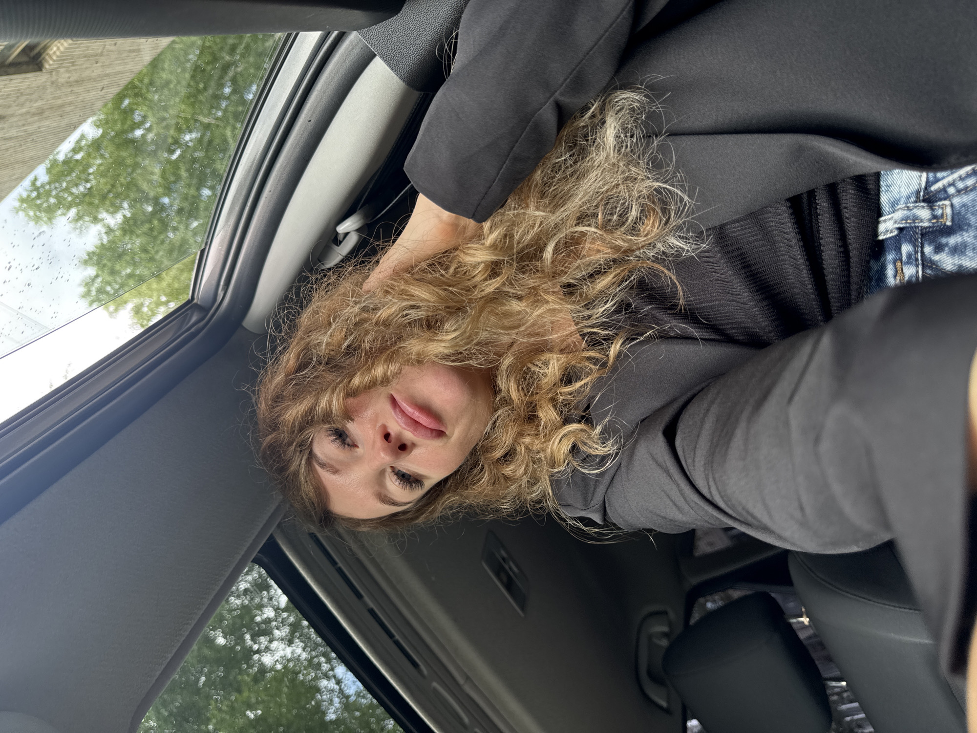 A young woman with long, curly blonde hair is sitting in a car, looking towards the camera. She is wearing a black blazer over a black ribbed top and blue jeans. The car has a sunroof that is open, allowing natural light to illuminate the scene. Outside the car, trees and a building are visible, suggesting an urban or suburban setting. The woman is touching her hair and appears to be taking a selfie. The overall lighting is bright, with the sunlight reflecting off the car's interior and the sunroof.