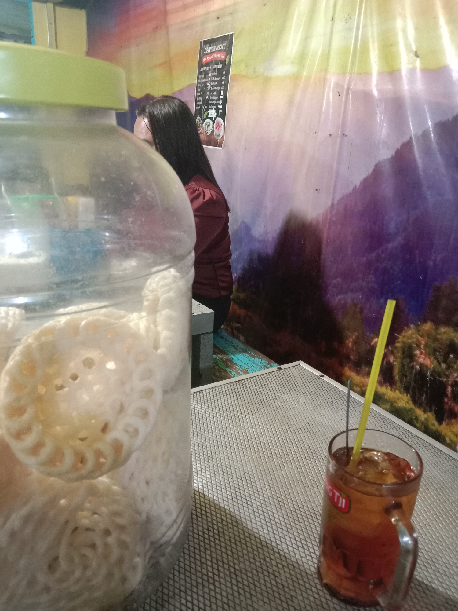 A close-up shot focuses on a clear glass jar filled with white, circular crackers, positioned in the foreground to the left. The jar has a light green lid. To the right of the jar, on a gray, textured surface, sits a glass mug filled with iced tea and a yellow straw. In the background, a woman with long dark hair and wearing a maroon top is partially visible, facing away from the camera. Behind her, a colorful mural depicts a mountainous landscape. A menu board is attached to the wall above the woman. The overall setting suggests an indoor eatery.