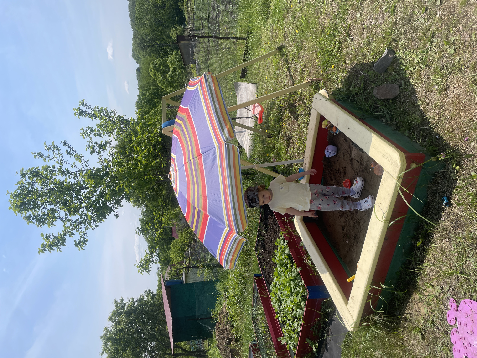 A young child stands in a sandbox, shaded by a multicolored striped umbrella. The sandbox is constructed from painted wooden boards in red, green, and yellow hues. The child wears a light-colored shirt, patterned pants, and white shoes.
The sandbox and child are situated in a grassy area, alongside a garden bed filled with leafy plants. Further back, there is a swing set and structures that appear to be a shed and fencing. The scene is outdoors, under a partially cloudy sky. The environment is indicative of a residential garden or yard.
While location details were requested, specific geolocation to Yasnoe, Russia, cannot be ascertained from the visual data alone. The visible characteristics are not distinctly indicative of a particular location.