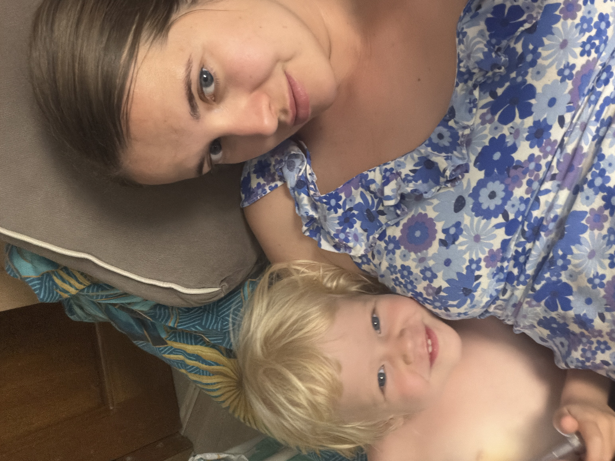 This is an overhead shot of a woman and a young child lying down together, likely taken in Sochi, Russia. The image is taken from a slightly upward angle, showing the woman's head and upper torso on the left, and the child's head and upper torso on the right, both looking towards the camera.
The woman has brown hair, blue eyes, and is wearing a blue, purple, and white floral patterned top with ruffled sleeves. She has a subtle smile on her face. Her head rests on a plain gray pillow, which in turn lies on a blue and yellow patterned fabric.
The child, with blonde hair and blue eyes, is lying next to the woman. They appear to be shirtless and have a light smile, looking directly at the camera. The same blue and yellow patterned fabric is visible beneath the child.
The background shows glimpses of what appears to be a wooden surface and possibly other bedding. The overall impression is one of warmth and closeness between the two individuals. There is no visible text in the image. The lighting suggests it might be daytime, possibly indoors.