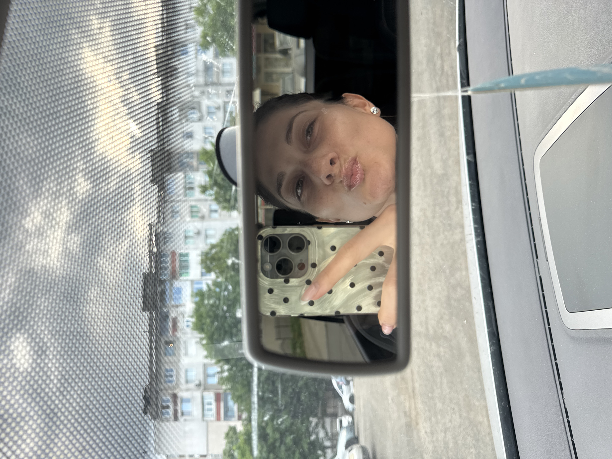 A person is visible in a car's rearview mirror, holding a phone. The reflection shows a woman with dark hair pulled back, wearing an earring. She is making a kissing face towards the camera. She is holding a phone with a light-colored case with black polka dots. The background, reflected through the mirror, shows a building and trees. The scene is viewed from inside the car, through the rearview mirror. Through the front window of the car, a building is visible, matching the architectural style and urban planning of Burgas, Bulgaria.