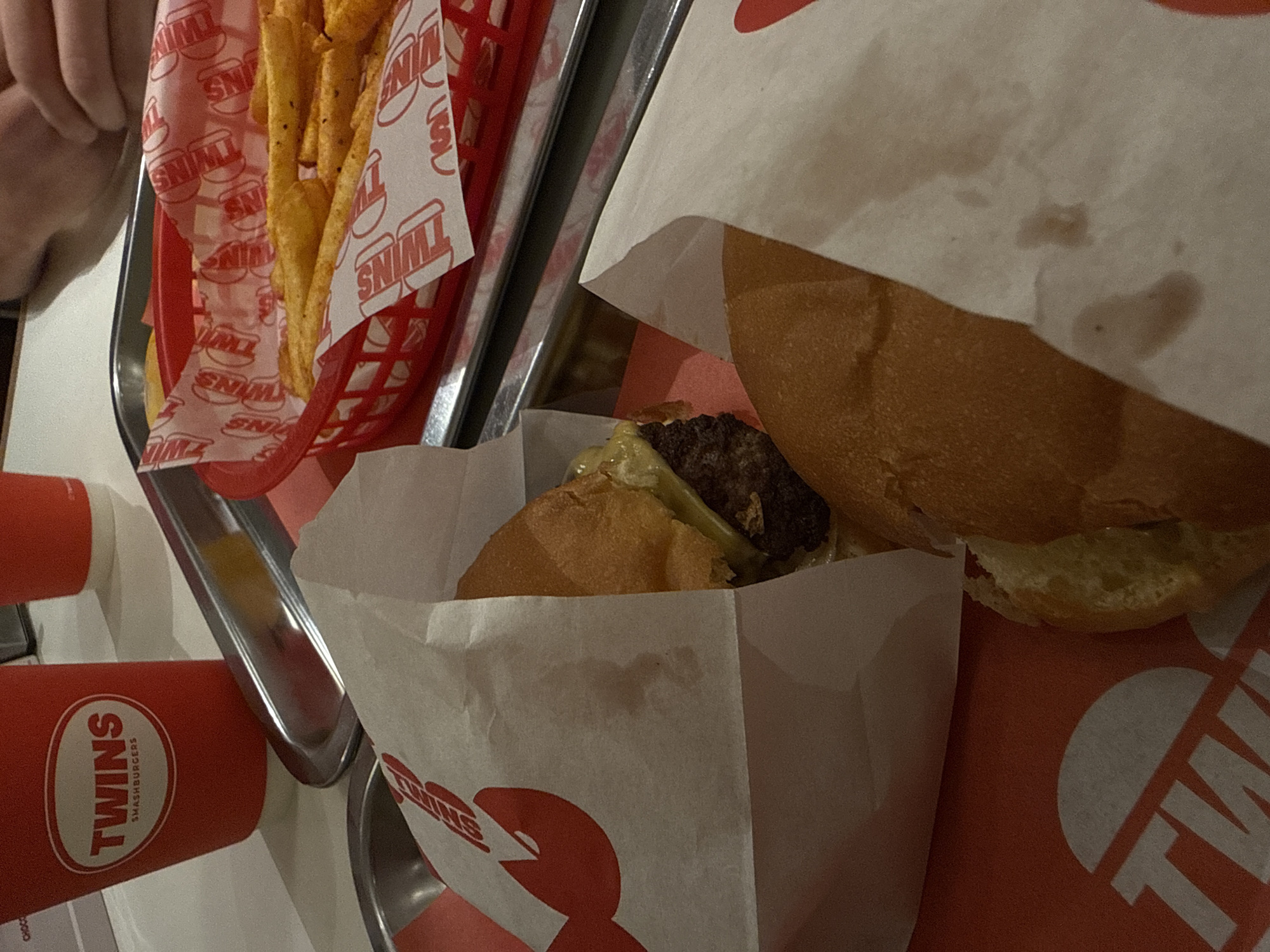 The image depicts a table with food and drink from "Twins Smashburgers" in Yerevan, Armenia. A red plastic basket contains french fries wrapped in branded paper. Two hamburgers are partially visible in paper bags, also branded with "Twins". Two red cups with the restaurant's logo are present. A hand is visible in the upper left corner. The food and drink items are situated on a metal serving tray.