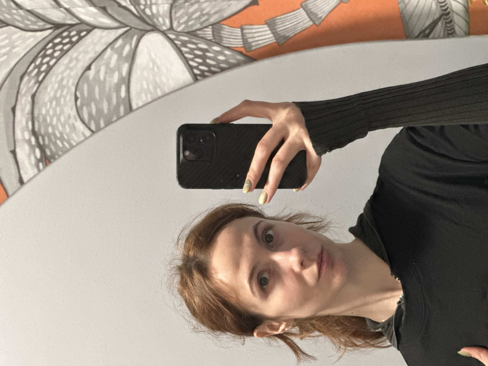 The image shows a woman taking a selfie in front of a mirror. The woman has light brown hair and is wearing a black long-sleeved shirt. She is holding a black smartphone in her right hand. Her fingernails are painted a pale yellow color.
The mirror reflects a portion of a wall with a decorative pattern in shades of gray, white, and orange. The wall appears to be painted white.
There is no explicit information to identify the location as Novi Sad, Serbia.