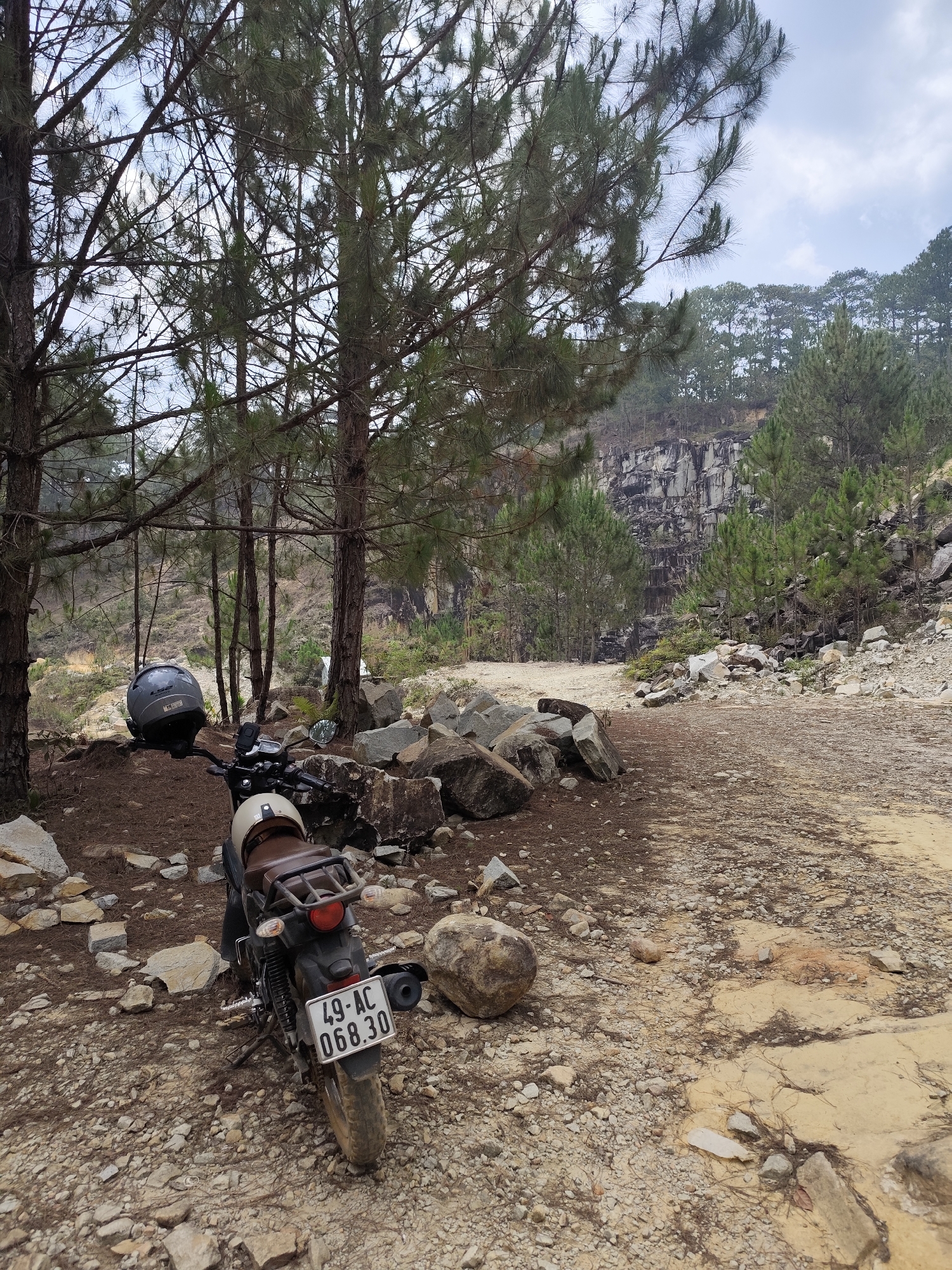 The image depicts a motorcycle parked on a gravel and rock-strewn area in a forest environment. The motorcycle, bearing a license plate "49-AC 068.30," is positioned near a cluster of trees. A helmet is placed on the motorcycle's handlebars. The surrounding area is composed of loose rocks, pebbles, and dirt. In the background, there is a cliff or rock face with vegetation growing on it. Based on visual cues, the location is Lang Biang, Da Lat, Vietnam. The scene appears to be a stop along a rugged or off-road trail. The weather conditions are overcast.