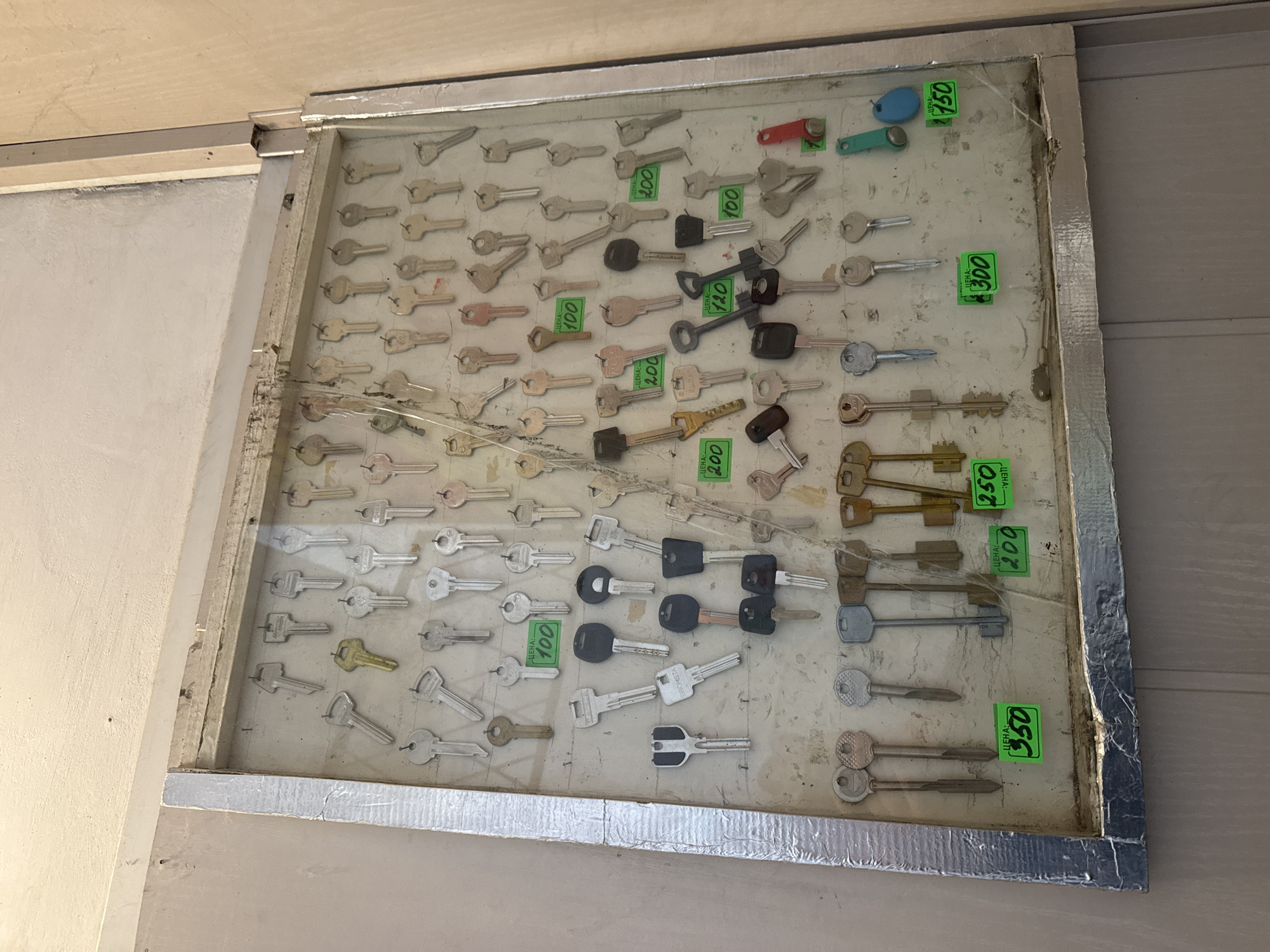 The image presents a display of various keys fixed on a white background, arranged within a silver-colored frame. Green price tags are affixed beside some of the keys. The prices marked on these tags include 100, 120, 150, 200, 250, 300, and 350, with the word "ЦЕНА:" and the word "BRONSTA" printed above some of the prices. The keys are of diverse types, some with black plastic heads, and others with metallic finishes. The frame appears old, with visible wear and tear. The background suggests that the display is mounted on a wall, possibly inside a shop or a service area. The city of Chaikovsky, Russia, is specified as the location of the photograph.