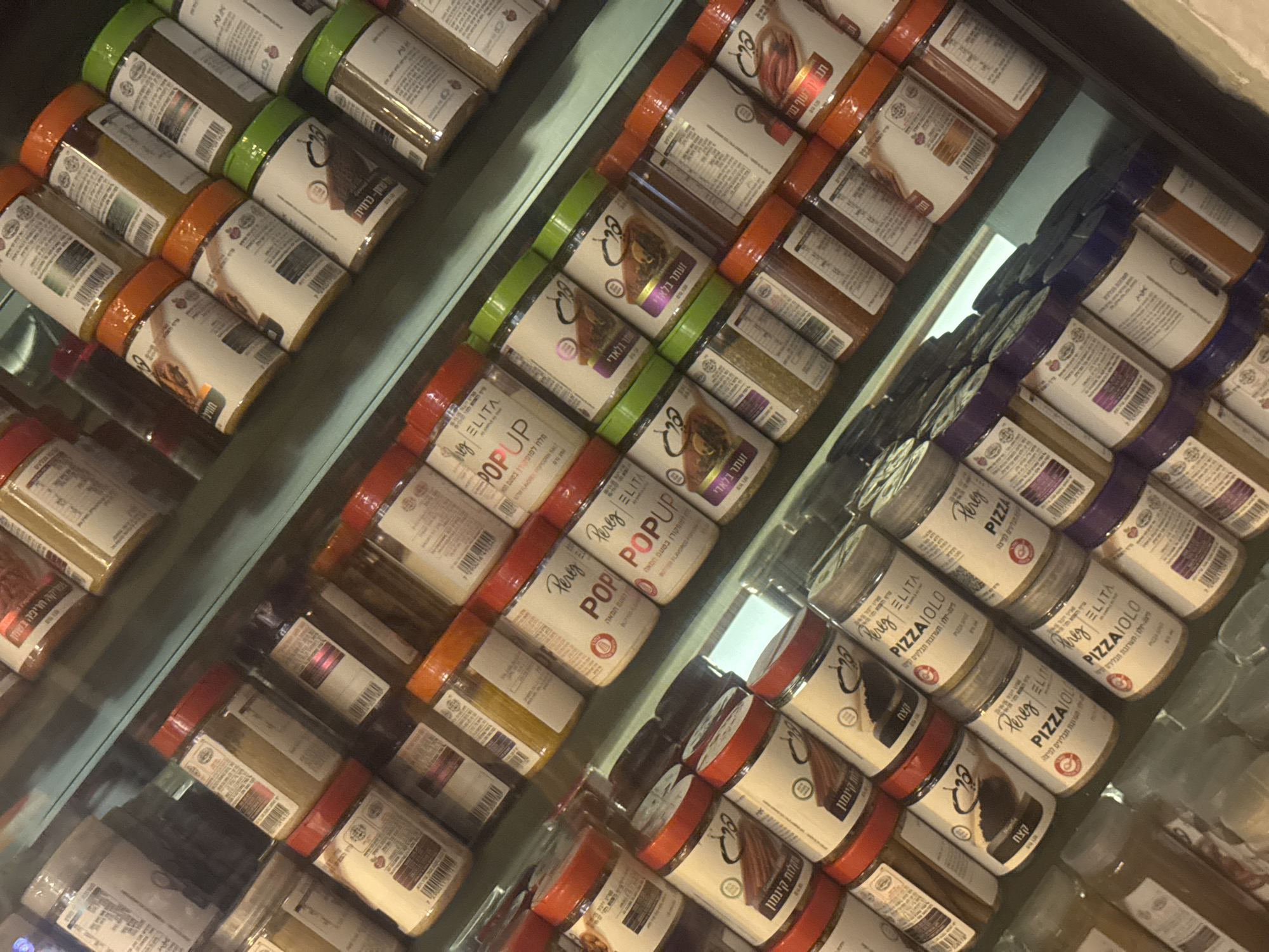 The image shows shelves stocked with jars of spices, specifically blends for popcorn and pizza. The brand "Pereg ELITA" is visible on many of the jars. Labels indicate flavors like "Butter Flavored Popcorn" and "Pizza" along with ingredients such as "Za'atar" and "Nigella Seed". The text on the labels is in Hebrew. The scene appears to be in a retail setting, likely a specialty food store or supermarket. The store is located in Ramat Gan, Israel.