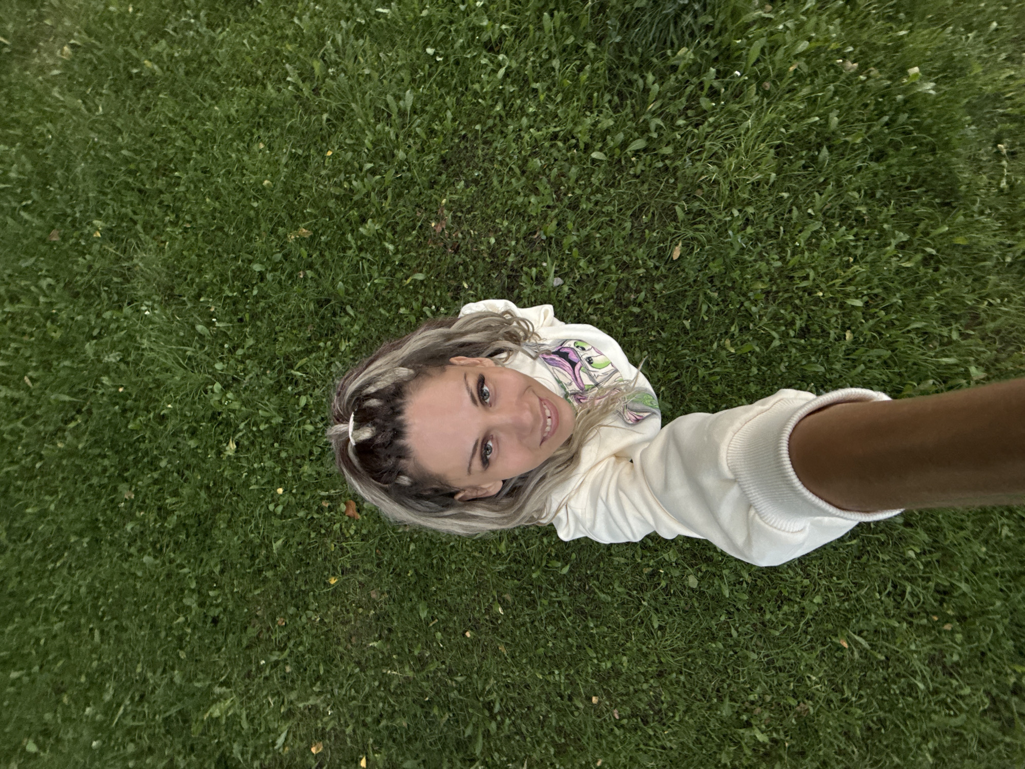 A high-angle, full-body shot shows a woman lying on a grassy field in Rusanovo, Russia. The woman is positioned in the center of the frame, looking up at the camera with a smile. She has blonde hair with streaks of gray and is wearing a light-colored hooded sweatshirt. The background is a lush green lawn, suggesting it is daytime and the weather is pleasant. The woman's arm is extended towards the camera, holding what appears to be a selfie stick or the camera itself. The scene evokes a sense of casual relaxation and enjoyment of the outdoors. No visible text is present in the image.