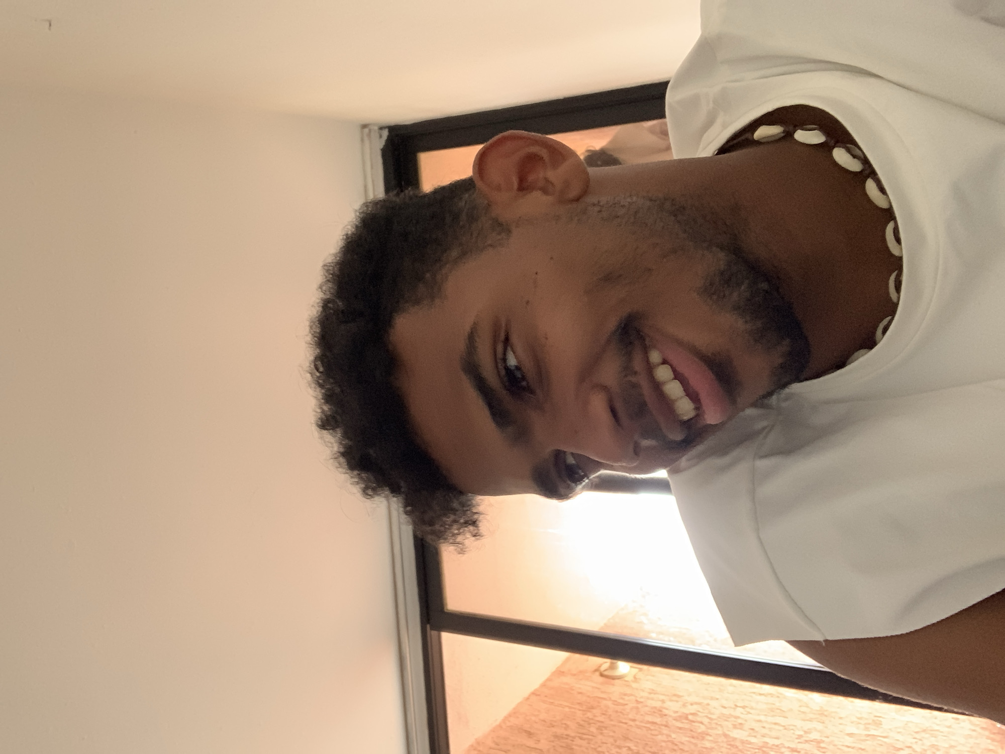 A close-up, portrait-style image captures a young man from Porlamar, Venezuela, smiling broadly with his mouth open, revealing his teeth and tongue. He has dark, curly hair and a light beard or stubble. He is wearing a white collared shirt or hoodie, and a distinctive necklace made of white cowrie shells is visible around his neck. The setting appears to be indoors, possibly near a window, with warm, bright light from outside filtering through. The light suggests either sunrise or sunset, bathing the background in a soft, orange glow, visible through dark window frames. The man's expression is one of joy or playfulness. There is no visible text in the image.