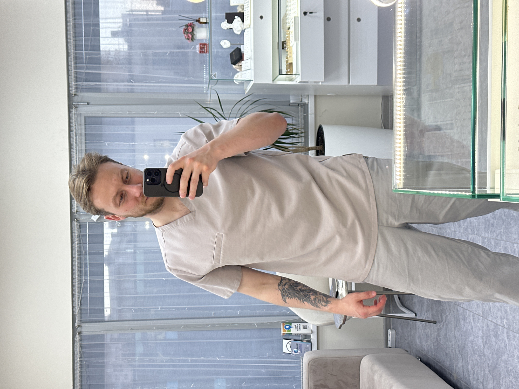 This image, rotated 90 degrees counter-clockwise, depicts a man taking a mirror selfie in what appears to be a commercial establishment.
The subject is a fair-skinned man with short, light brown hair and stubble, wearing a light-colored (beige or light grey) scrub-style uniform consisting of a V-neck top with a small chest pocket and matching pants. He holds a dark-colored smartphone with a triple camera system in his right hand, pointing it towards himself to capture the reflection. A prominent tattoo, appearing to be a detailed animal (possibly a wolf or similar creature) design, covers a significant portion of his left forearm. He has a slight, relaxed smile.
The setting is an indoor space, likely a retail store, clinic, or office, characterized by a modern and clean aesthetic. In the background, large windows are visible, covered with sheer, light-colored curtains or blinds. To the man's right, there are multiple white display cabinets and glass display cases, some featuring internal strip lighting. These cases contain various small items, possibly jewelry or decorative objects. On a shelf above the display cases, a plant in a white pot, a diffuser with sticks, and a small vase with pink flowers can be seen. On the far right, part of a light-colored sofa or bench is visible. The floor is covered with light grey tiles or linoleum.
The overall atmosphere is bright, suggesting either ample natural light filtering through the windows or strong artificial illumination, particularly from the lit display cases. There is some blurry, illegible text visible on small labels near the man's left leg, but no clear words can be deciphered.