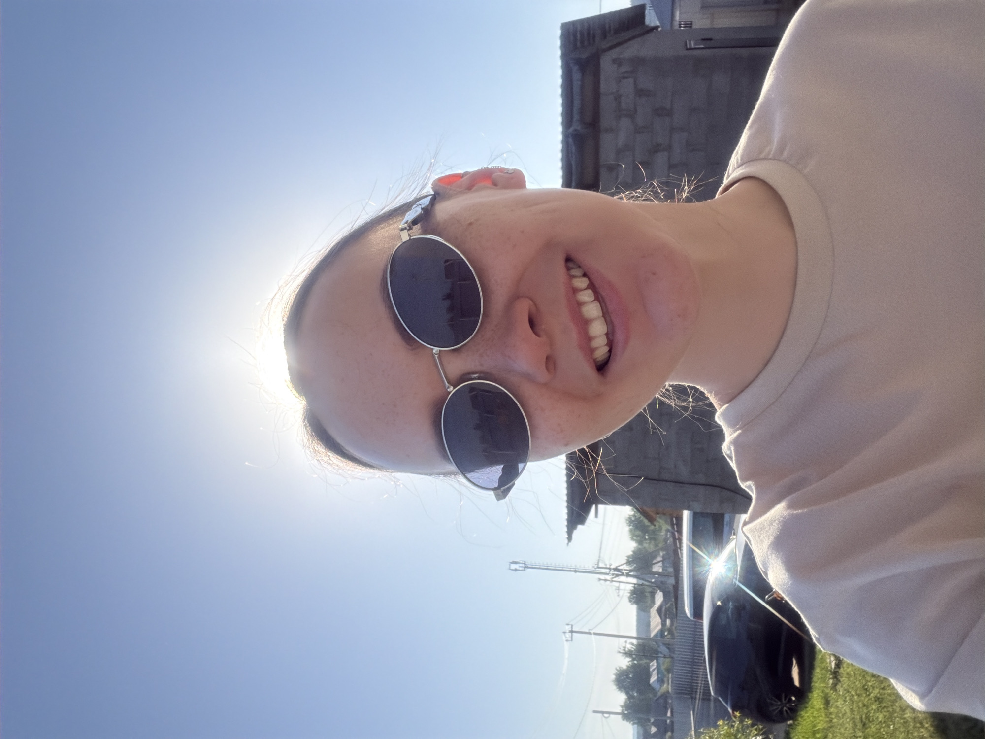 A young, light-skinned woman is facing the camera with a smiling expression while wearing round sunglasses with dark lenses and a silver frame. The background reveals a sunny, outdoor scene with a clear blue sky partially obscured by the brightness of the sun in the upper left corner. In the distance, there are buildings constructed of gray brick and wood, interspersed with utility poles and trees. A section of a dark-colored vehicle, likely a car, is visible to the right. The location is Chaikovsky, Russia based on available data. The focus is on the woman's face and upper body, with the environment serving as a backdrop.