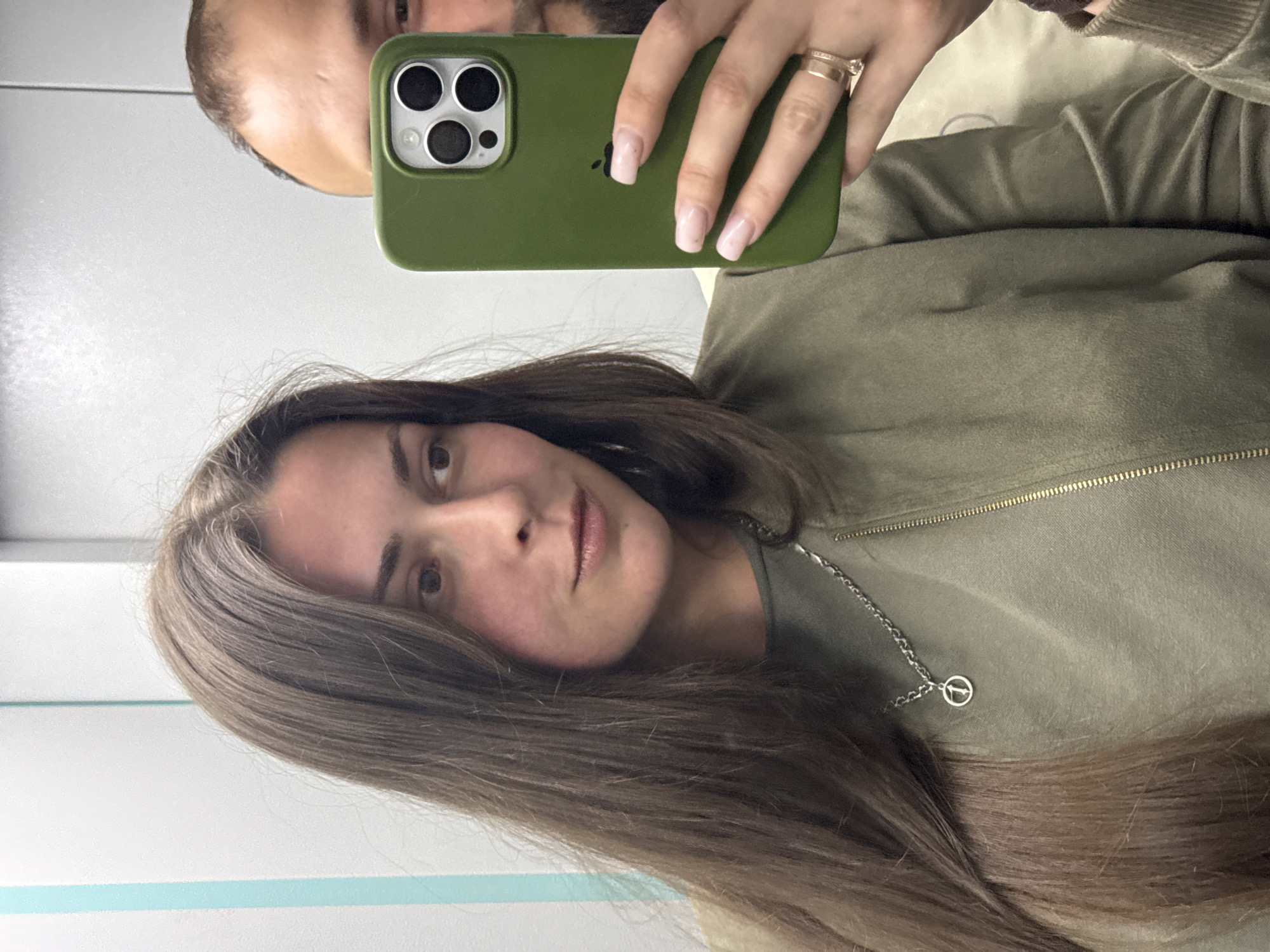 A woman and a man are reflected in a mirror, taking a selfie. The woman, in the foreground, has long, brown hair and is wearing a green jacket over a green top. She is also wearing a silver necklace with a circular pendant. The man, in the background, is partially visible, holding a green iPhone in his right hand. He has short, dark hair and is wearing a light-colored shirt. The background appears to be a wall with a vertical blue stripe. The scene is indoors, possibly in an elevator or a hallway.